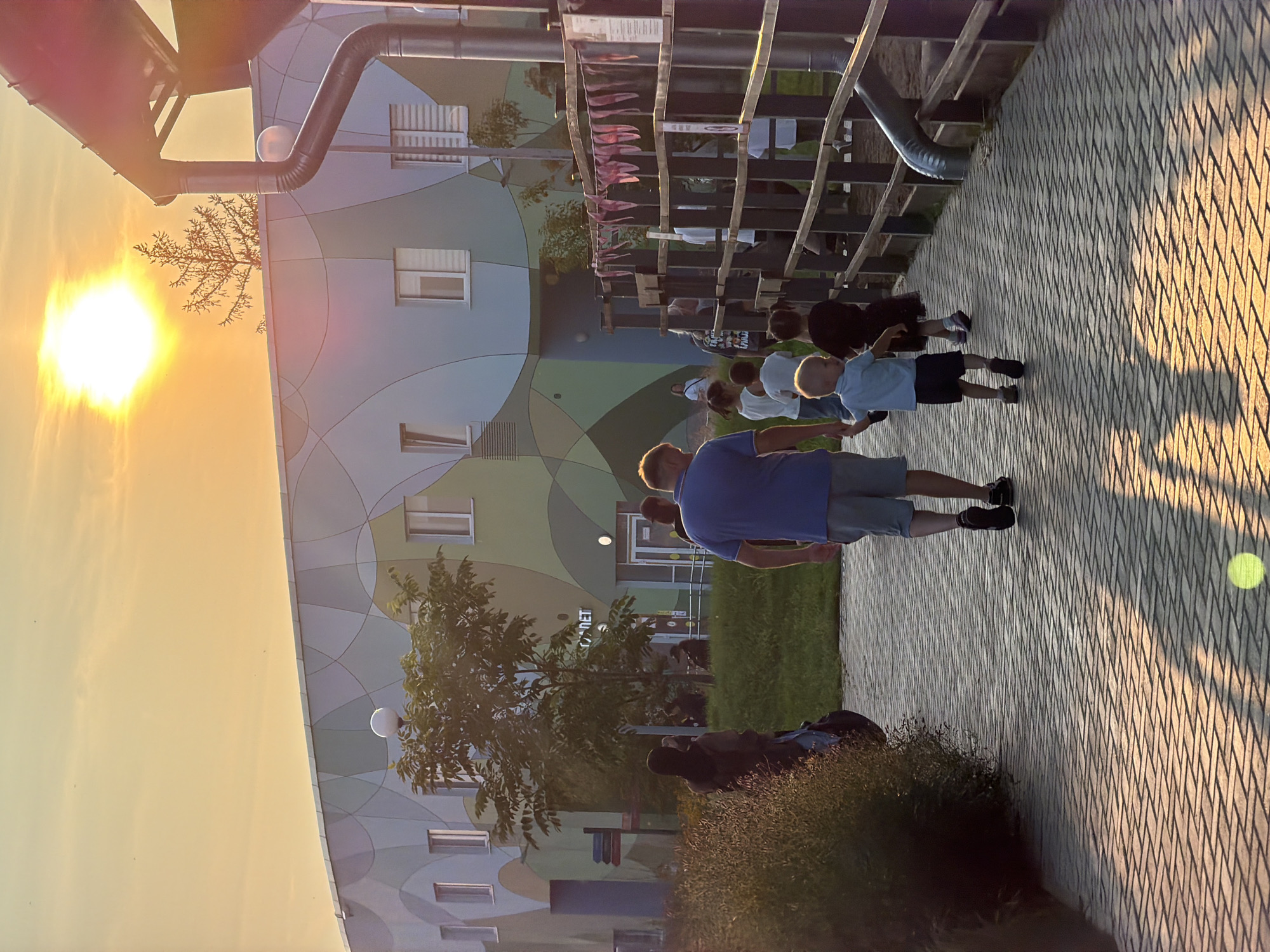 This media file depicts an outdoor scene in Perm, Russia, during what appears to be a late afternoon or early evening, given the warm, golden sunlight casting long shadows. The sun is prominently visible in the upper left, creating a bright glow.
The main subjects are a group of people, including adults and children, standing or walking on a paved pathway. The pathway is laid with patterned tiles that create a diamond-like effect. To the left of the pathway is a grassy area and a building with a distinctive, colorful facade. The building's exterior is decorated with large, curved shapes in shades of light blue, green, and beige, with visible windows. A large metal pipe runs diagonally across the upper part of the image, above the building. A wooden structure, possibly a fence or part of a playground, is also present, adorned with small pink flags.
The people are casually dressed and appear to be engaged in some form of outdoor activity or gathering. Some are facing away from the camera, while others are in profile. A child in a blue shirt and dark shorts is walking with his arms outstretched. An adult in a blue t-shirt and light shorts is standing closest to the camera. The overall mood seems relaxed and pleasant, consistent with a warm, sunny day.
Visible text includes the word "ДЕТ" on the building, which is likely part of a larger word like "ДЕТСКИЙ" (children's) or "ДЕТСКИЙ САД" (kindergarten), suggesting the building might be a children's facility. There is also some unreadable text on signs attached to the wooden structure.