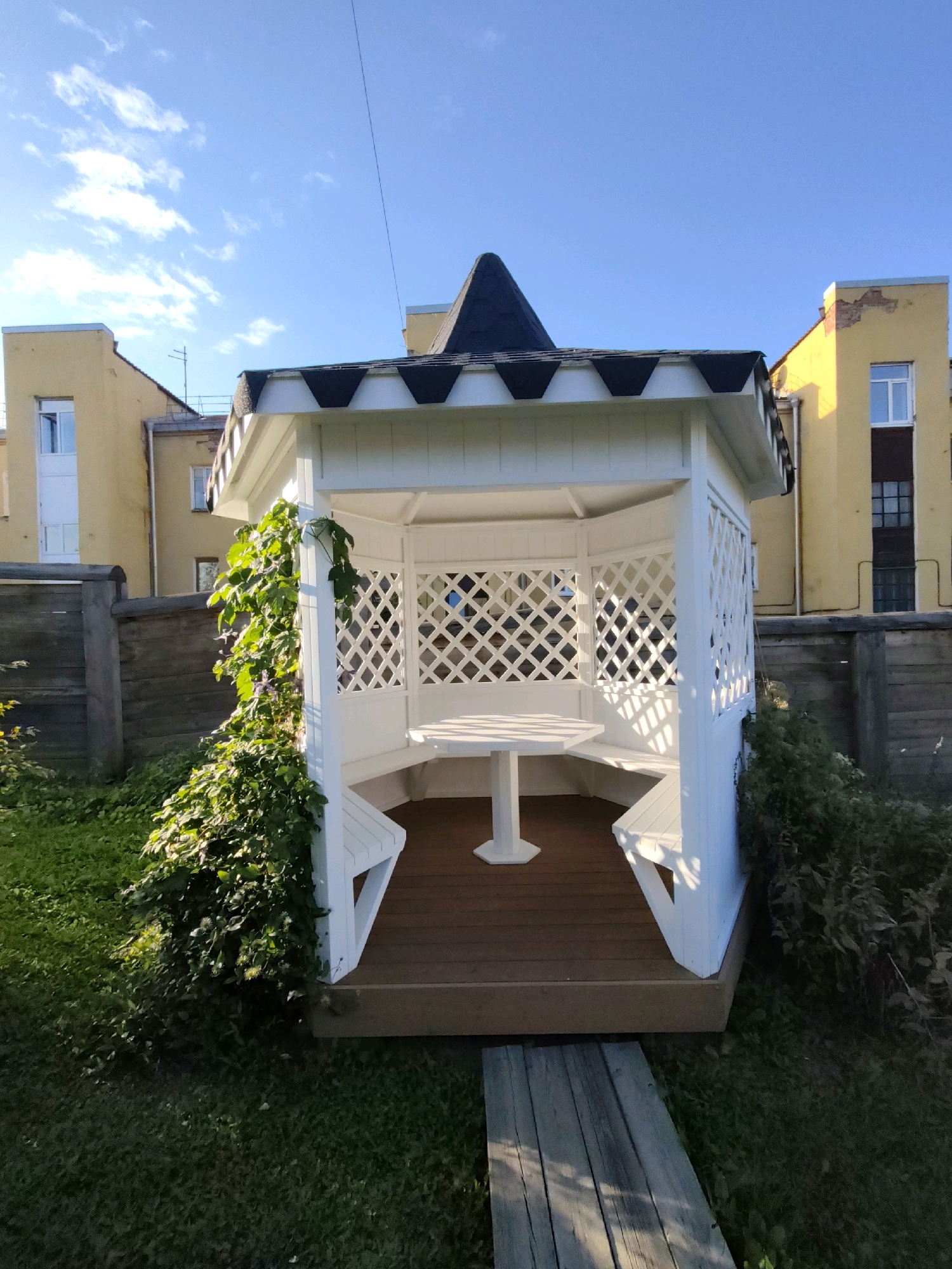 The image shows a white wooden gazebo with a brown wooden floor, situated in a grassy yard. The gazebo has a hexagonal shape and features lattice panels on its sides. A white table and benches are visible inside. The gazebo is surrounded by green foliage, and a wooden fence runs along the background. Behind the fence, there are yellow buildings with windows. The sky is clear and blue with a few white clouds. There are no people or animals in the image. The scene appears to be in a residential area, possibly a backyard or a public park. The architectural style of the yellow buildings suggests an older, possibly Soviet-era construction. Without further information or context, the exact location in Irkutsk, Russia, cannot be determined from the image alone.
