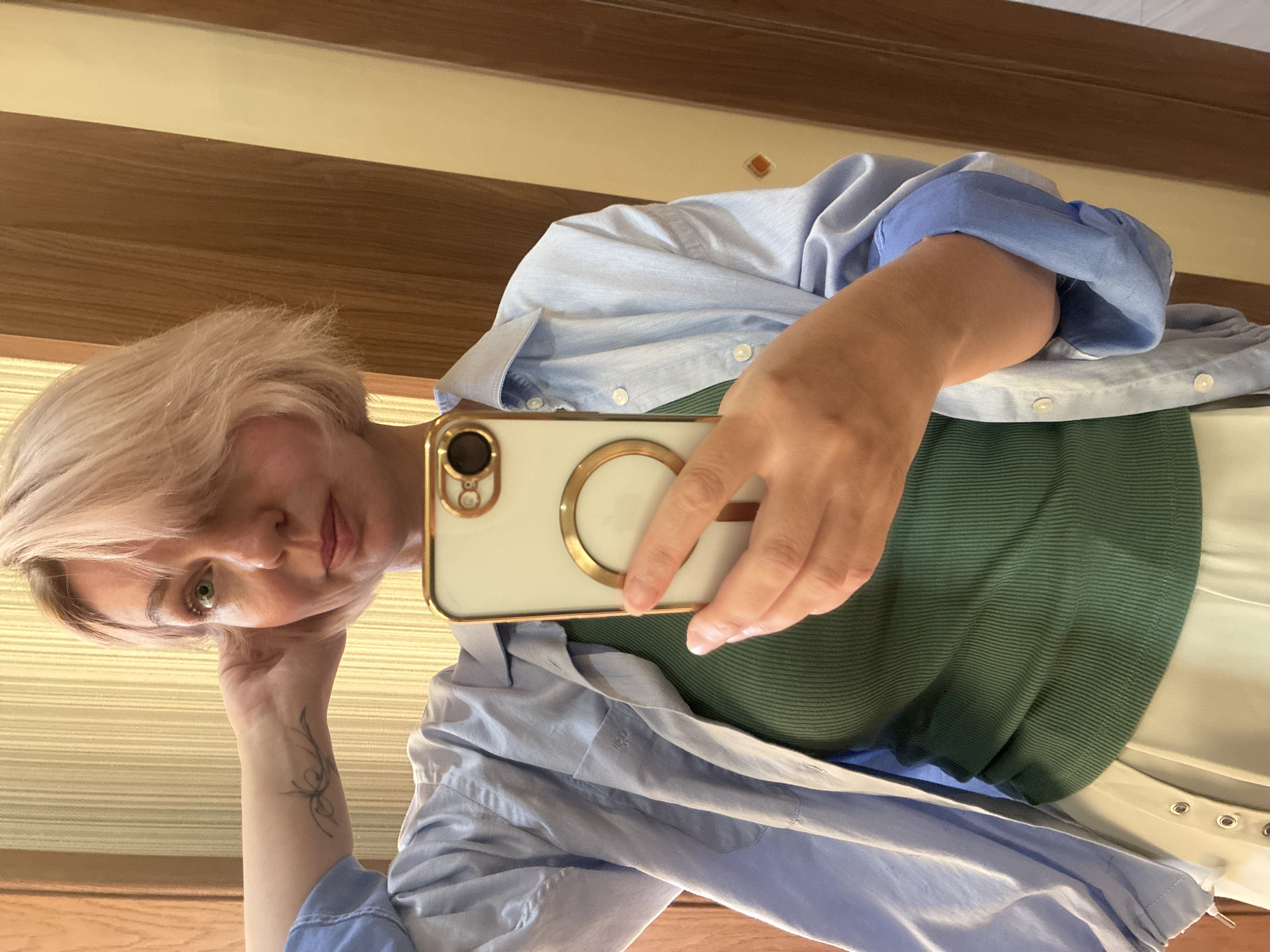 In the city of Sochi, Russia, a fair-skinned female with short, light pink-blonde hair is captured in a mirror selfie. Her gaze is directed slightly upwards and to the right of the camera. She wears a light blue, unbuttoned, long-sleeved collared shirt over a fitted, dark green ribbed tank top. A light-colored garment, possibly white or light beige pants or a skirt with visible metal eyelets on the waistband, is seen below the tank top. Her left hand is raised towards the left side of her head, with a small, stylized tattoo visible on her left inner wrist. Her right hand holds a light-colored smartphone with a gold frame and camera accents, featuring a prominent circular gold accessory on its back, pointed towards the viewer. The background, visible in the mirror's reflection, consists of vertical wooden paneling or doors on the right and a lighter, possibly striped wall or textured surface on the left.