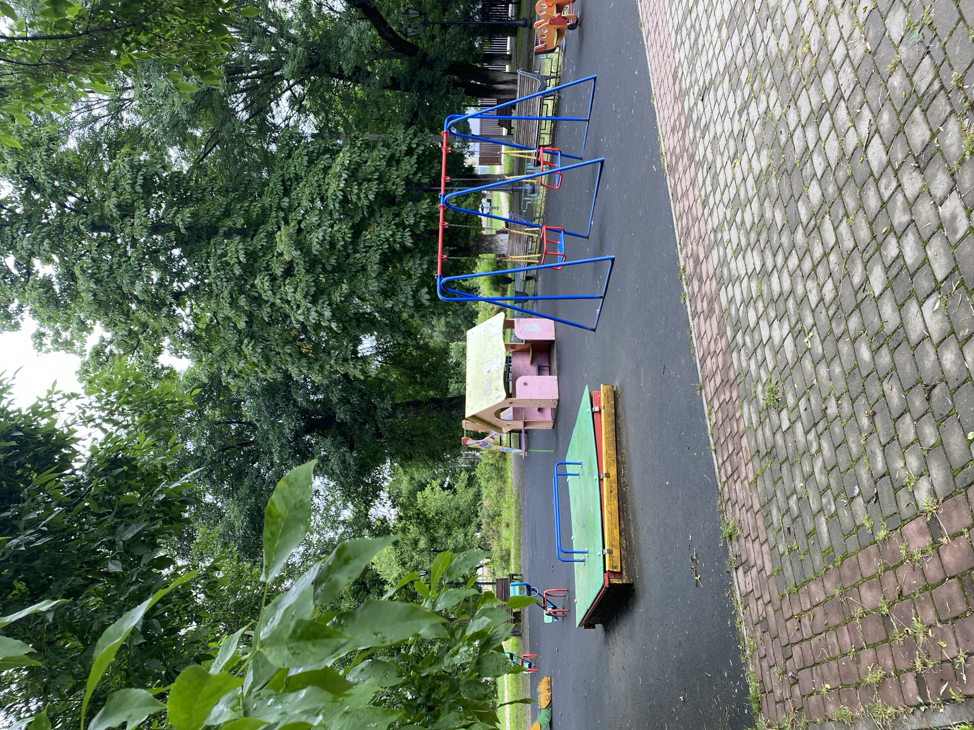 This is a photograph of a playground in the city of Zapadny, Russia. The playground is situated in a park-like setting with mature trees providing shade. The ground is covered in a dark, resilient surface, with a paved brick pathway bordering the area in the foreground.
The playground equipment includes a sandpit with a green lid and blue railings, a pink playhouse with a green roof, and a blue swing set with two red swings. In the background, there are several park benches and what appears to be a small amusement ride with cartoon-like characters. The foliage is lush and green, suggesting it is summertime or late spring. The overall scene depicts an empty, tranquil playground on an overcast day.