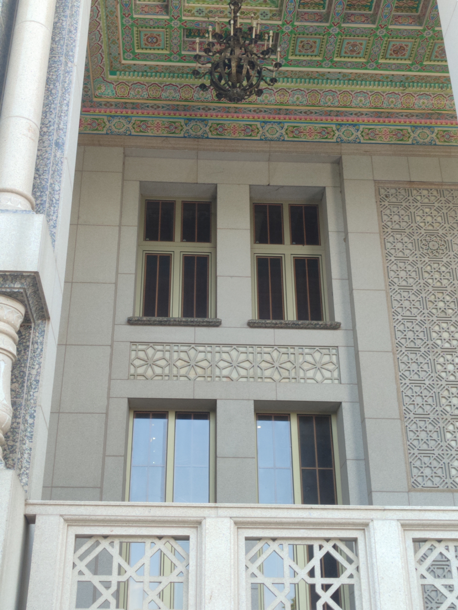 The image shows the facade of a building in Dushanbe, Tajikistan, likely an administrative or public building, featuring a combination of classical and Islamic architectural elements. The upper part of the facade is adorned with a richly decorated, multi-colored ceiling or soffit, featuring geometric patterns and intricate floral motifs in shades of green, red, blue, and gold. A large, ornate chandelier hangs from this ceiling. Below the decorative ceiling, there are two sets of large, vertically oriented windows with dark-tinted panes, divided into multiple sections. The windows are framed by light-colored stone. Between the upper and lower windows, there is a decorative stone frieze with repeating star-shaped geometric patterns. The lower part of the visible facade includes a balcony or railing with a similar geometric stone lattice design, behind which more windows can be seen. The building's exterior walls are constructed from large, light-colored stone blocks. To the left of the windows, a large, fluted column with blue and white patterned tiles adds to the building's ornate appearance. The architectural style suggests a building of historical or cultural significance within Dushanbe.