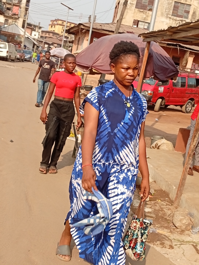A woman in a blue and white patterned dress walks along a street. She carries a colorful floral purse and a blue-patterned cloth. Behind her, another person wears a red cropped top and dark trousers, with a man in a black t-shirt and jeans visible further back. A red van and other vehicles are partially visible amidst buildings and street structures. The scene suggests an outdoor market or busy street in a developing area.