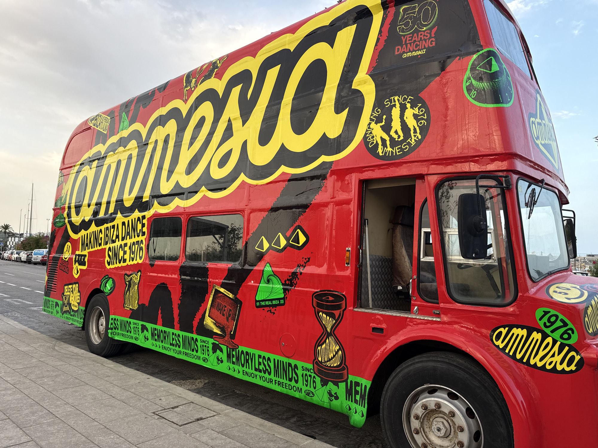 A red double-decker bus with the word "amnesia" emblazoned on its side in large yellow lettering is parked on a sidewalk in Ibiza, Spain. The bus is covered in various graphics and text, including "50 YEARS DANCING AMNESIA," "MAKING IBIZA DANCE SINCE 1976," and "MEMORYLESS MINDS SINCE 1976 ENJOY YOUR FREEDOM." The bus features a black and red design, with yellow accents and green circular emblems. The scene appears to be a street in Ibiza, possibly near the port, given the presence of cars and buildings in the background. The sky is overcast.