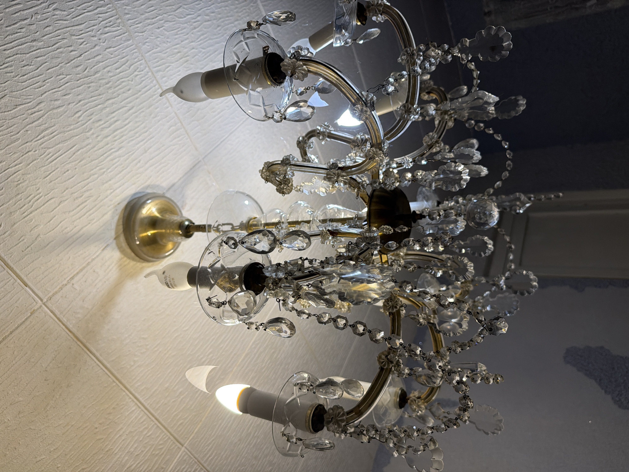 An ornate brass chandelier with numerous crystal pendants hangs from a textured white ceiling. The chandelier has at least four candle-shaped light bulbs, two of which are illuminated, casting a warm glow. The ceiling appears to be made of acoustic tiles. The background consists of a dark blue wall with a subtle textured pattern, and a glimpse of a white doorframe is visible on the right side of the image. No people or animals are present in the scene. The lighting suggests an indoor setting, possibly a residential or hospitality space, with the photograph taken from a low angle looking upwards. The scene is located in the city of Sochi, Russia.