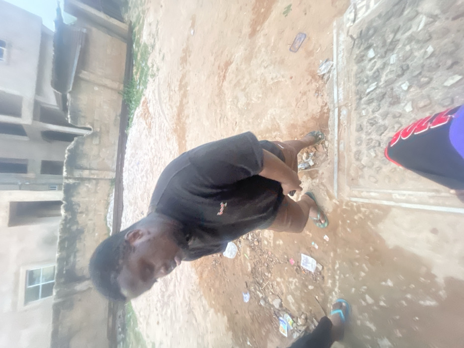 The image, when rotated clockwise by 90 degrees, displays an outdoor scene from a slightly elevated perspective, focusing on individuals on a damp, unpaved path.
In the center-left, a person with dark skin and short dark hair is seen, facing slightly away from the camera and looking downwards. They are dressed in a plain black short-sleeved t-shirt, which features a small, light-colored logo (resembling a stylized 'Y' or similar character) on the left chest. They are also wearing dark shorts and light green flip-flops.
To the right side of the image, the lower body of at least two other individuals is partially visible. In the immediate foreground on the right, a foot wearing a light blue flip-flop is visible. Further back and slightly to the right, another person's lower leg and foot are seen. This person is wearing a black shoe with distinctive red accents or patterns, and a purple lower garment (possibly a skirt or trousers). The word "SOUL" in white letters with red outlines is clearly visible on the side of this black shoe.
The ground beneath the individuals is unpaved, composed of sandy earth or dirt, and appears significantly damp, with visible puddles or wet patches, suggesting recent rain or poor drainage. Scattered litter, including various plastic bags and wrappers, is strewn across the ground. A dark rectangular object, possibly a mobile phone, lies on the ground near the center of the frame. A narrow, light-colored concrete strip, resembling a curb or divider, runs vertically through the mid-ground, separating the muddy path from a rougher, textured paved area to its right.
In the background to the left, several multi-story buildings are visible. These structures appear to be made of concrete or block, with some walls looking unfinished or exposed. Multiple windows are visible on the building facades, indicating a residential or commercial setting within an urban or semi-urban environment.
The lighting is bright and diffused, suggesting it is daytime, possibly under an overcast sky, as no harsh shadows are apparent. The overall impression is of people navigating a somewhat neglected outdoor area with basic infrastructure.