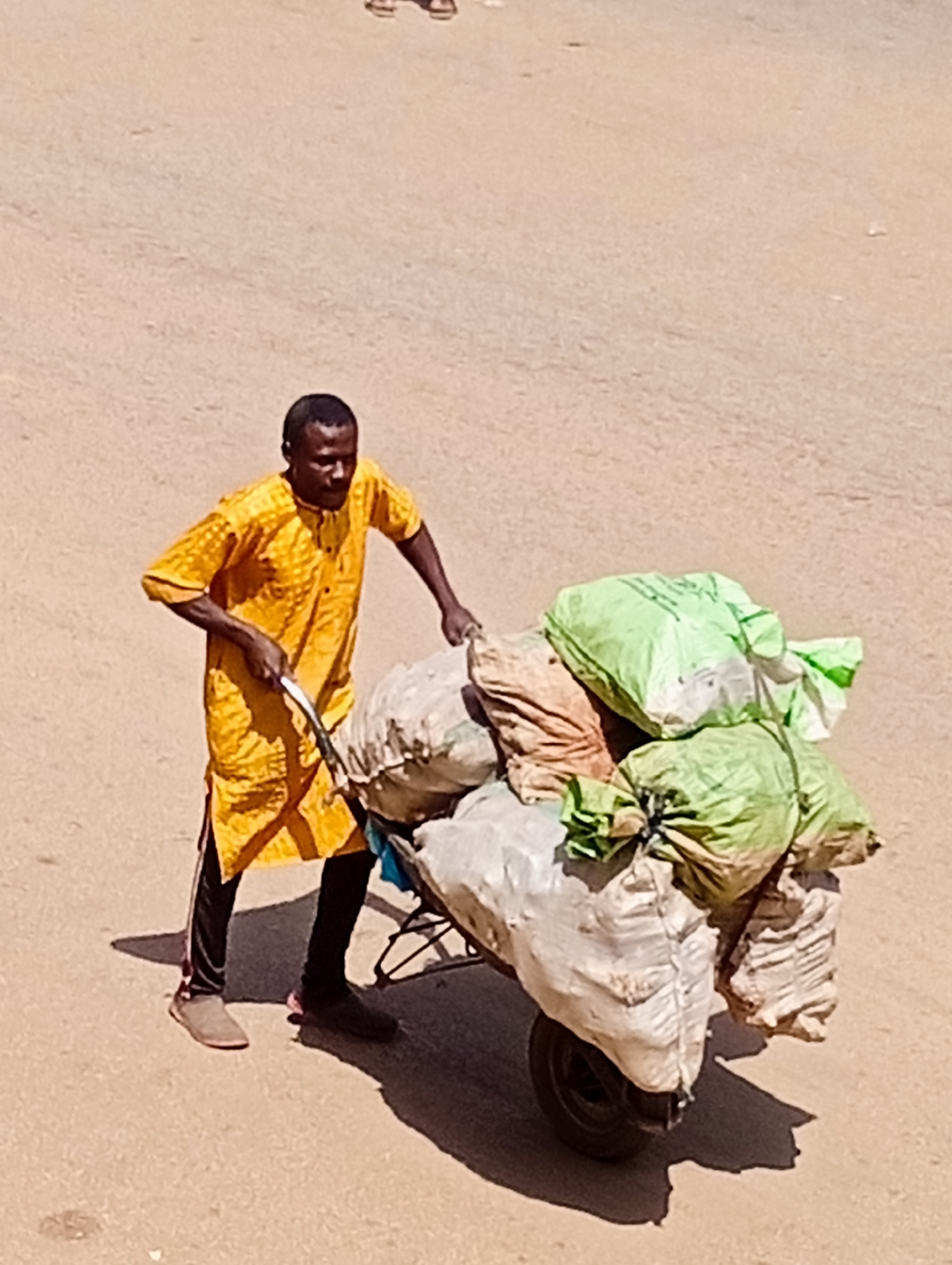 A Black man, wearing a yellow traditional garment and black pants, pushes a two-wheeled cart laden with several large sacks. The man is facing right, with his left hand on the cart's handle. The sacks on the cart are of various colors and sizes, including light green, off-white, and beige. The cart has a black wheel visible on the right side. The ground is a light brown, sandy or dusty surface, indicative of a road or unpaved area. The scene appears to be outdoors, possibly in a marketplace or street within the city of Bukuru, Nigeria. Sunlight casts shadows on the ground.