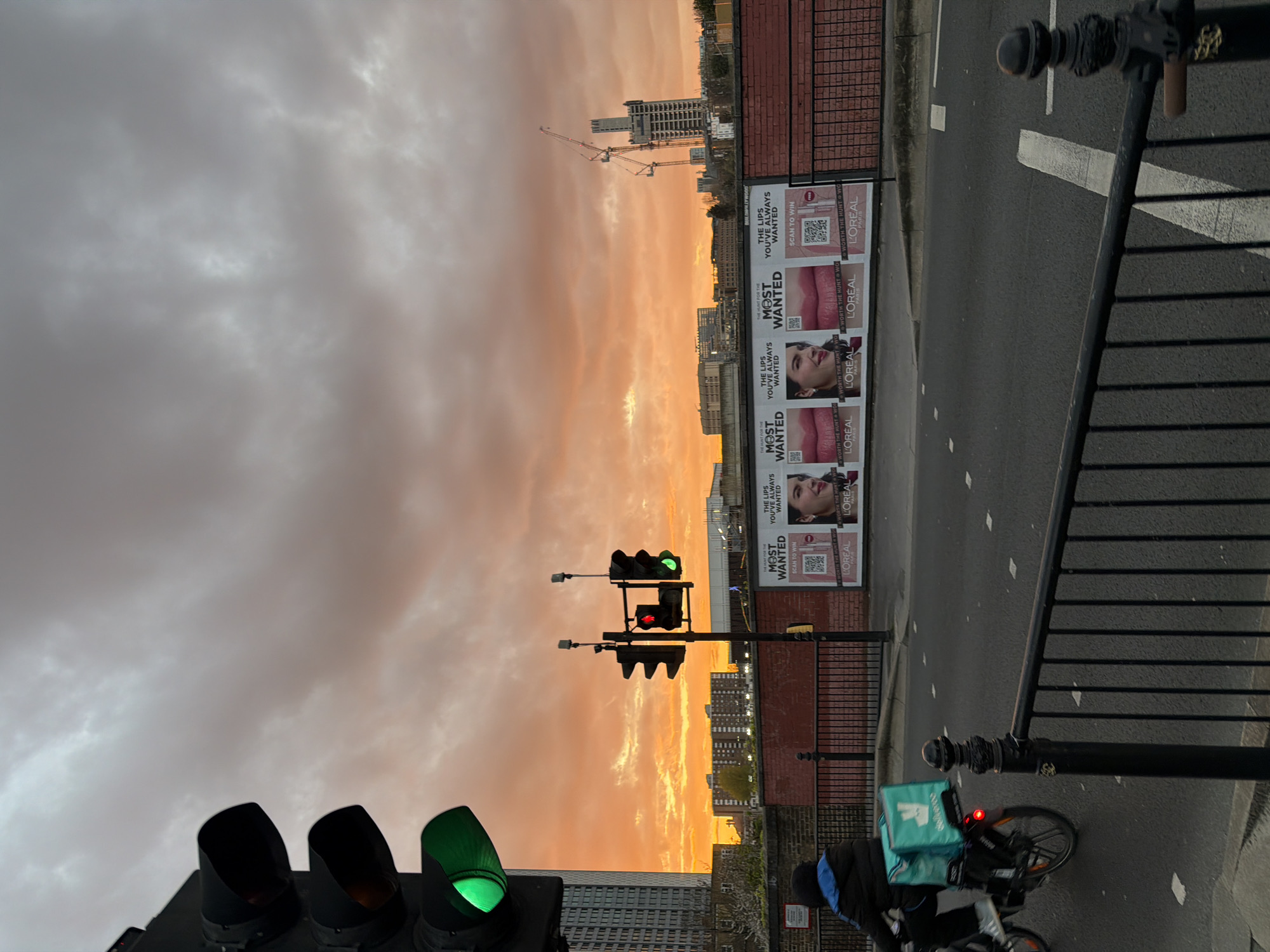 Here is a detailed description of the image:
The image captures an urban scene in London, United Kingdom, under a striking sunset sky filled with orange-tinted clouds. A road runs through the frame, alongside a black metal railing. On one side of the road, there are multiple advertising posters for L'Oreal, featuring the slogan "THE LIPS YOU'VE ALWAYS WANTED". The sky dominates the background, with city buildings visible in the distance, including a construction site with cranes. In the foreground, there are traffic lights displaying green and red signals. A cyclist with a Deliveroo delivery bag is present, partially obscured by the railing. The overall scene portrays a juxtaposition of city life and the natural beauty of a sunset.