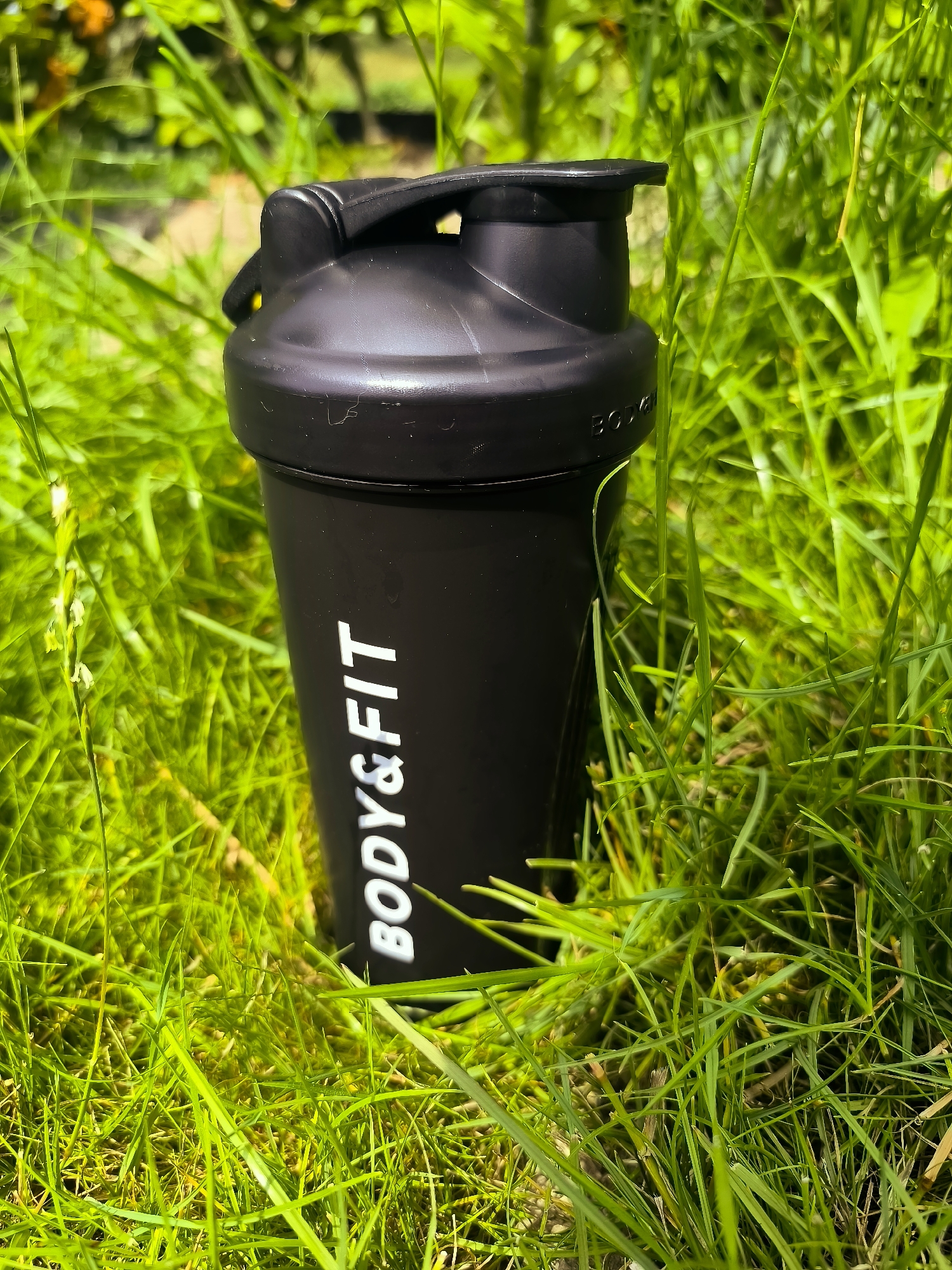A black shaker bottle with white text that reads "BODY&FIT" is placed upright in lush green grass. The bottle has a black lid with a flip-top closure. The background consists of more grass and out-of-focus foliage, suggesting an outdoor setting. The lighting indicates it is daytime, likely sunny due to the bright green of the grass and the contrast. There is no indication of Almere, Netherlands, in the image.