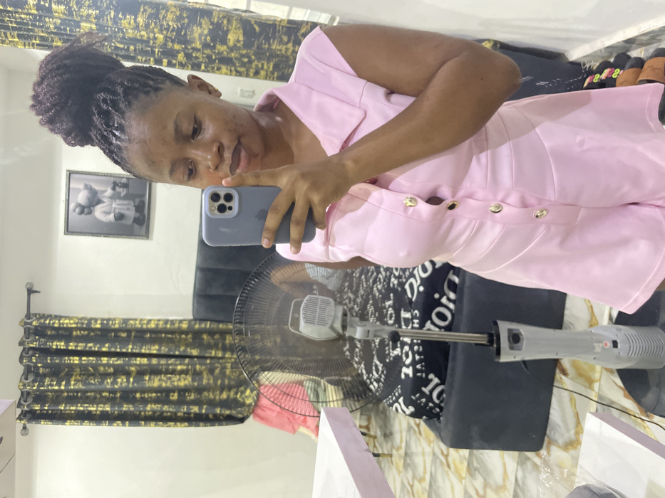 A female subject is depicted taking a mirror selfie indoors in Okpanam, Nigeria. The subject, a woman with brown skin and dark, textured hair styled in a high bun, is wearing a short-sleeved, collared pink romper with gold buttons down the front. She holds a light gray smartphone with multiple rear cameras to her face, capturing her reflection.
The background, visible in the mirror's reflection, includes a dark gray upholstered headboard or sofa. A black and white patterned sheet, possibly featuring the text "DIOR," covers a bed or mattress behind this furniture. A gray standing floor fan is positioned in the midground. On the wall to the right, a framed artwork displays two cartoon-like figures in a grayscale palette. Dark curtains with a gold abstract pattern are visible on the left side of the background. The floor features light-colored patterned tiles. A light pink surface, likely a table, is partially visible on the far left. Items resembling sandals are partially visible on the floor at the bottom right.