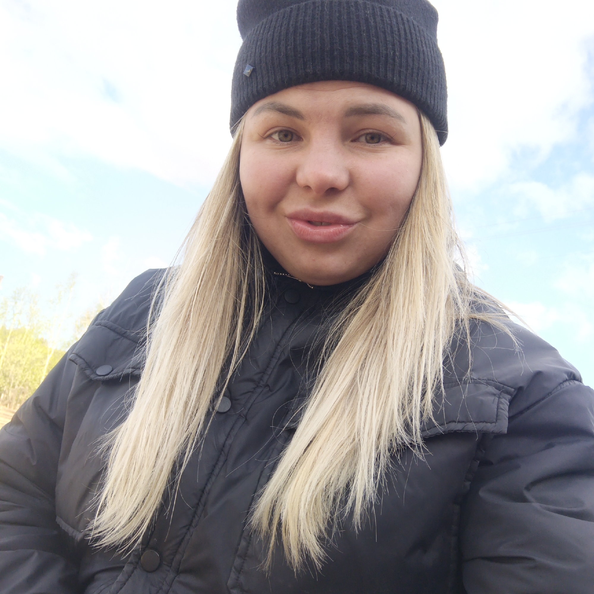 A woman is centered in the frame wearing a black beanie and black puffy jacket, with long blonde hair. The background is slightly out of focus and appears to be a pale blue sky with some clouds. Some vegetation is visible in the far background on the left. Location is Ваньки, Russia.