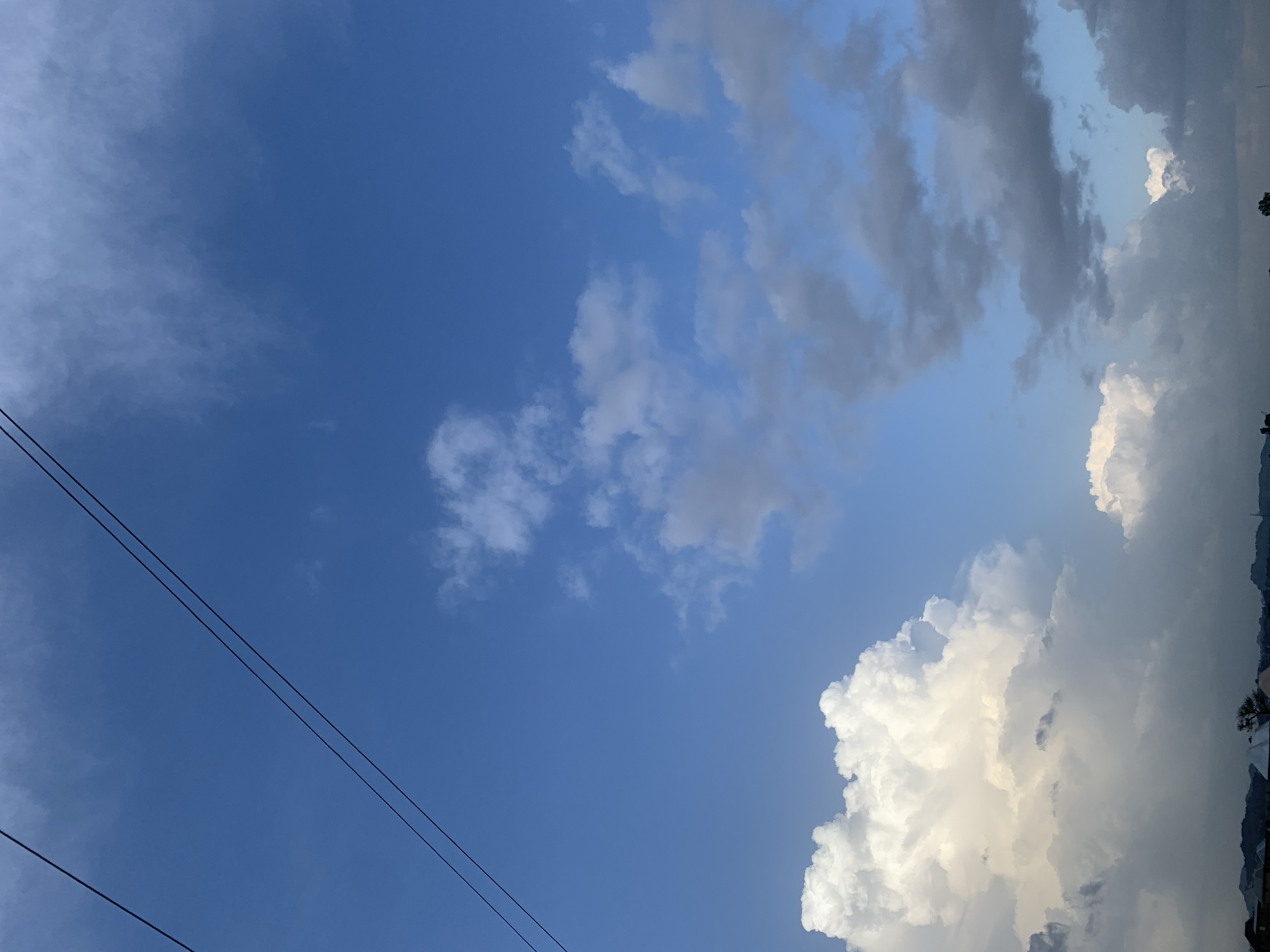 The image depicts a vast expanse of blue sky over Kuru Karama, Nigeria, with a dramatic display of clouds. Two parallel black wires stretch diagonally across the upper left quadrant of the frame, silhouetted against the bright sky. The clouds vary in form and color, ranging from bright white, puffy cumulus formations in the lower left to darker, more ominous gray clouds scattered throughout the mid-ground and right side. The lower edge of the image shows a dark, indistinct silhouette of a cityscape or rural landscape, with hints of buildings and trees, suggesting the geographical context of Kuru Karama. The lighting indicates it is either early morning or late afternoon, given the contrast between the illuminated white clouds and the shadowed areas. No people or specific actions are visible.