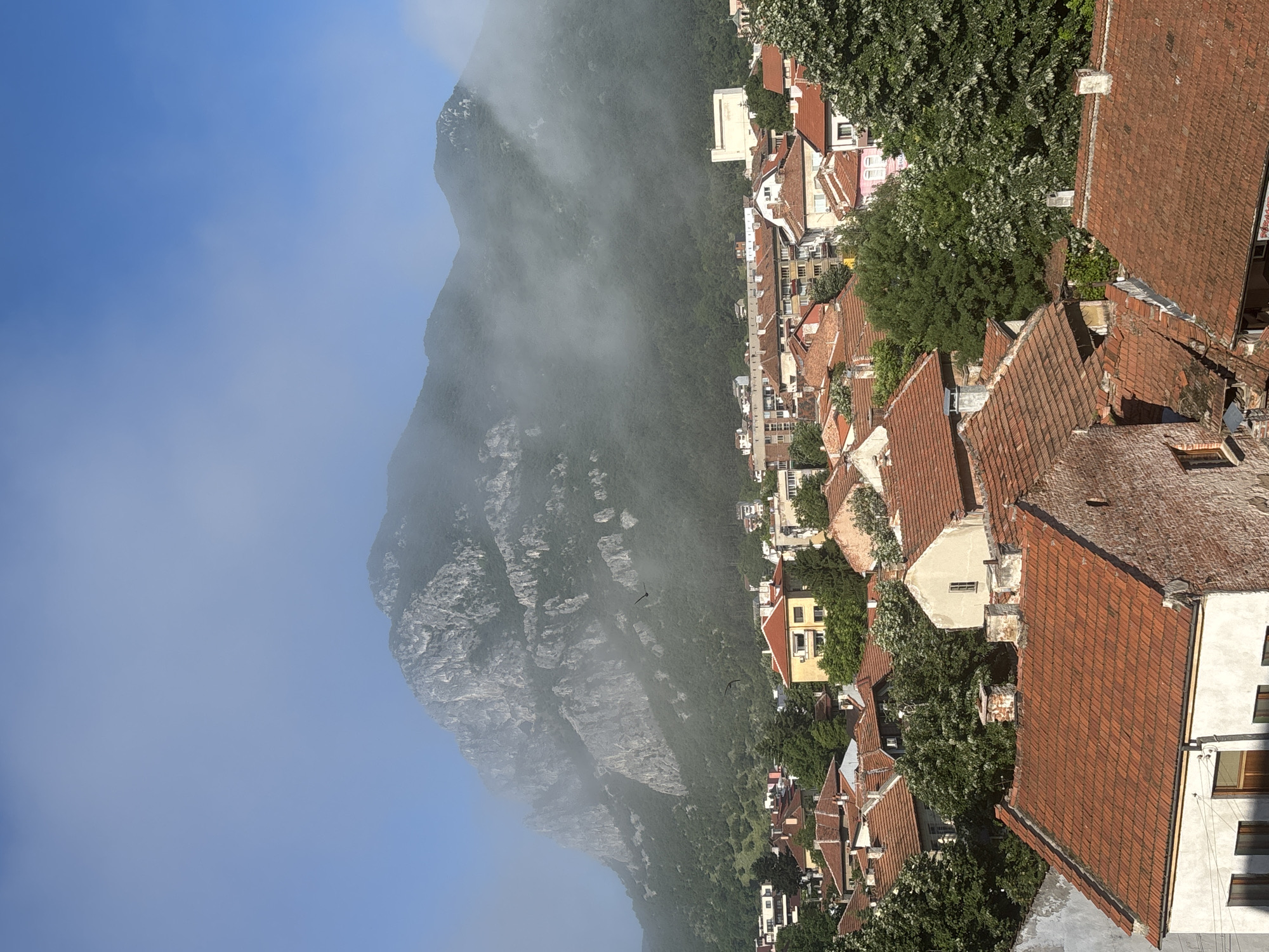 This landscape photograph captures a panoramic view of the town of Vratsa, Bulgaria, situated at the base of a majestic mountain range, the Vrachanski Balkan.
In the foreground and mid-ground, a dense collection of buildings with distinctive reddish-orange tiled roofs sprawls across the terrain. The architecture appears traditional, with many roofs showing age and character, suggesting a rich history. Green trees are abundant, interspersed among the houses, providing a lush contrast to the urban structures.
The formidable Vrachanski Balkan mountains dominate the background, their rugged, rocky slopes covered in dense green vegetation. A layer of mist or low-lying clouds partially shrouds the higher peaks and contours of the mountain, creating an atmospheric and somewhat mystical effect. Above the clearer sections of the mountain, a bright, clear blue sky is visible. Two dark silhouettes of birds can be seen in flight against the mountainous backdrop.
The scene depicts a serene daytime setting, bathed in natural light. There are no specific human activities or individuals prominently featured, placing emphasis on the architectural and natural beauty of the Bulgarian landscape. The image evokes a sense of tranquility and the harmonious coexistence of the town with its grand natural surroundings. There is no visible text in the media file.