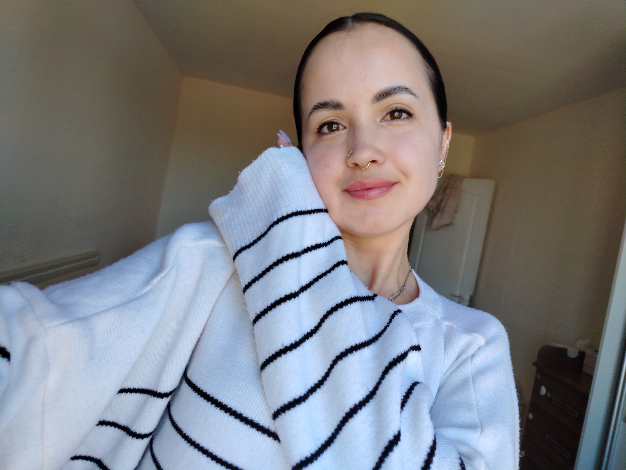 A woman is taking a selfie indoors. She is wearing a white sweater with black horizontal stripes and has dark hair pulled back from her face. She has pierced ears and a nose ring. Her right hand is near her face. The room is painted in neutral tones, and a door and wooden dresser are visible in the background. The location is not in Noviy, Russia; the image shows an interior setting that does not provide any information about its location, and thus I cannot confirm it is in Noviy, Russia.