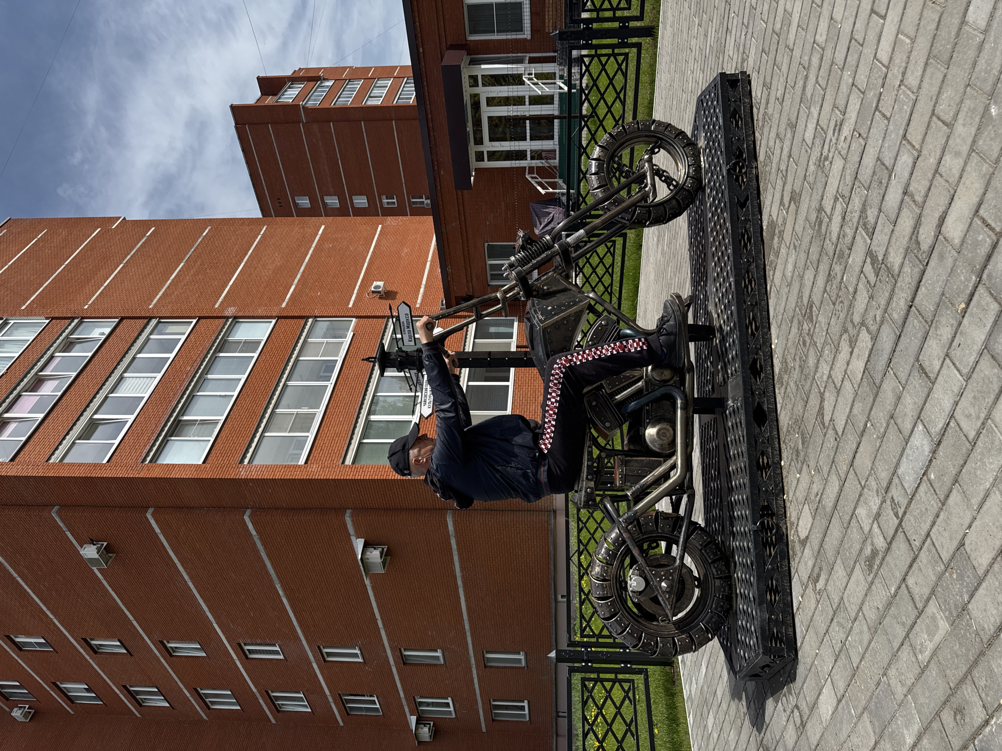 A person wearing a black baseball cap, black jacket, and black pants with red and white checkered stripes along the side seams sits on a metal motorcycle sculpture. The motorcycle is dark gray and appears to be made of welded metal parts. It is mounted on a black metal platform. The person is holding onto the handlebars as if riding the motorcycle.
The scene is set outdoors on a paved area of gray bricks. In the background, there is a tall brick building with multiple windows arranged in rows. To the right of the motorcycle, there is a short black metal fence and a patch of green grass.
The location is Chaikovsky, Russia. A street sign in the background indicates the name of a street, written in Russian Cyrillic.