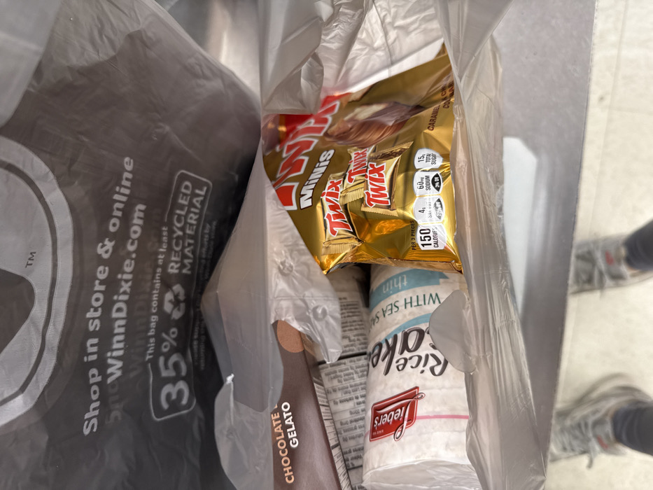 A bag of groceries contains a bag of Twix Mini chocolate candy, a package of Wieber's thin rice cakes with sea salt, and chocolate gelato. The plastic grocery bag has "Shop in store & online SnoWinnDixie.com" printed on it, indicating the store where the items were purchased. The bag also specifies it contains at least 35% recycled material. The nutritional information on the Twix bag shows 150 calories per 3 pieces.