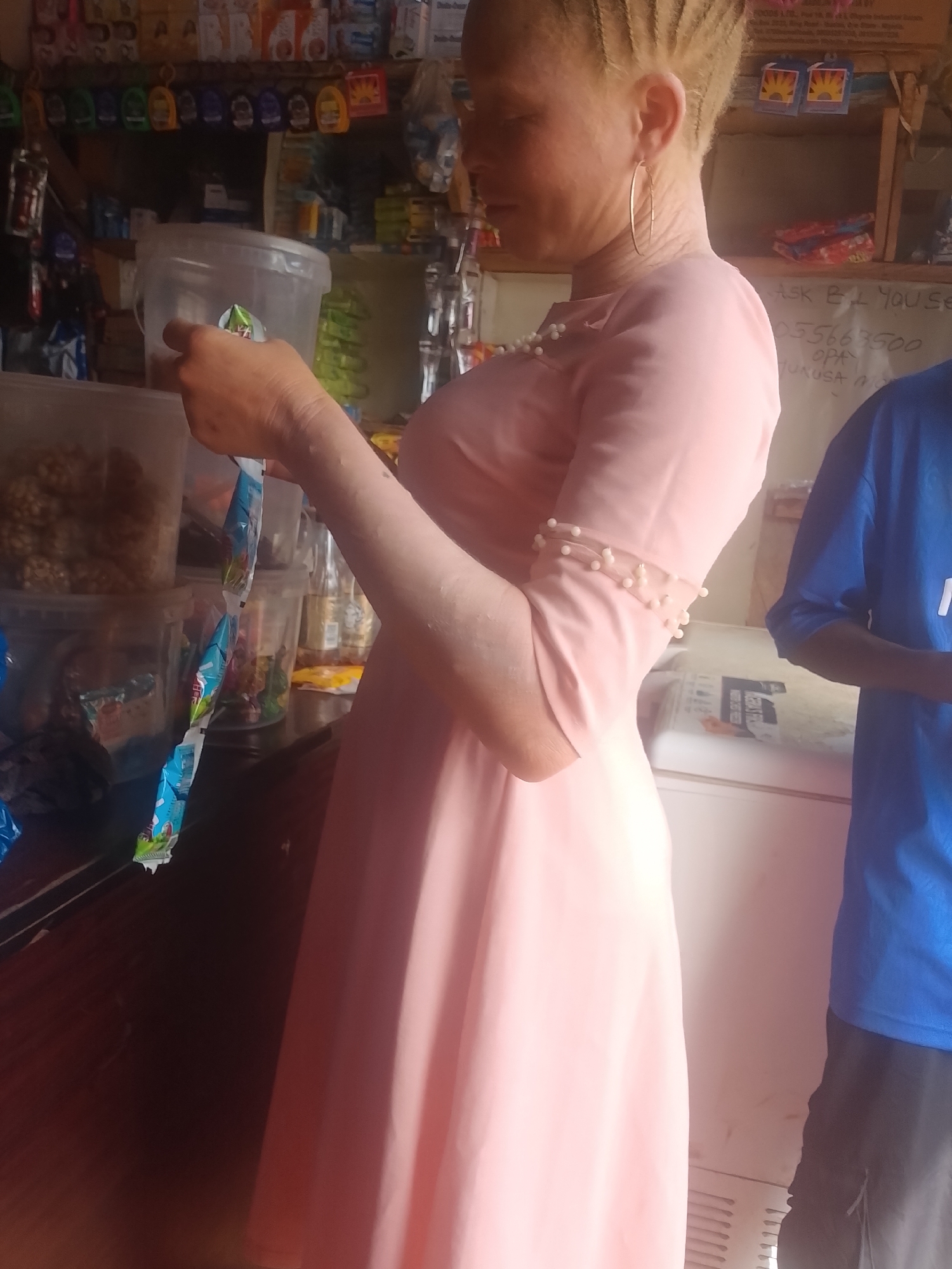 The image shows a fair-skinned person with braided hair, wearing a pink dress with pearl-like accents on the sleeves. They are holding what appears to be several small packets of blue and white packaged goods. The setting appears to be a small store or shop, with shelves stocked with various goods in the background. In the lower right of the frame is another person wearing a blue shirt and dark pants. The text "ASK B.I. YOU SEE... 0556683500... OPA..." can be seen in the background. Based on the text, this location could be Ejule, Nigeria. A white refrigerator is also visible behind the person in the pink dress.