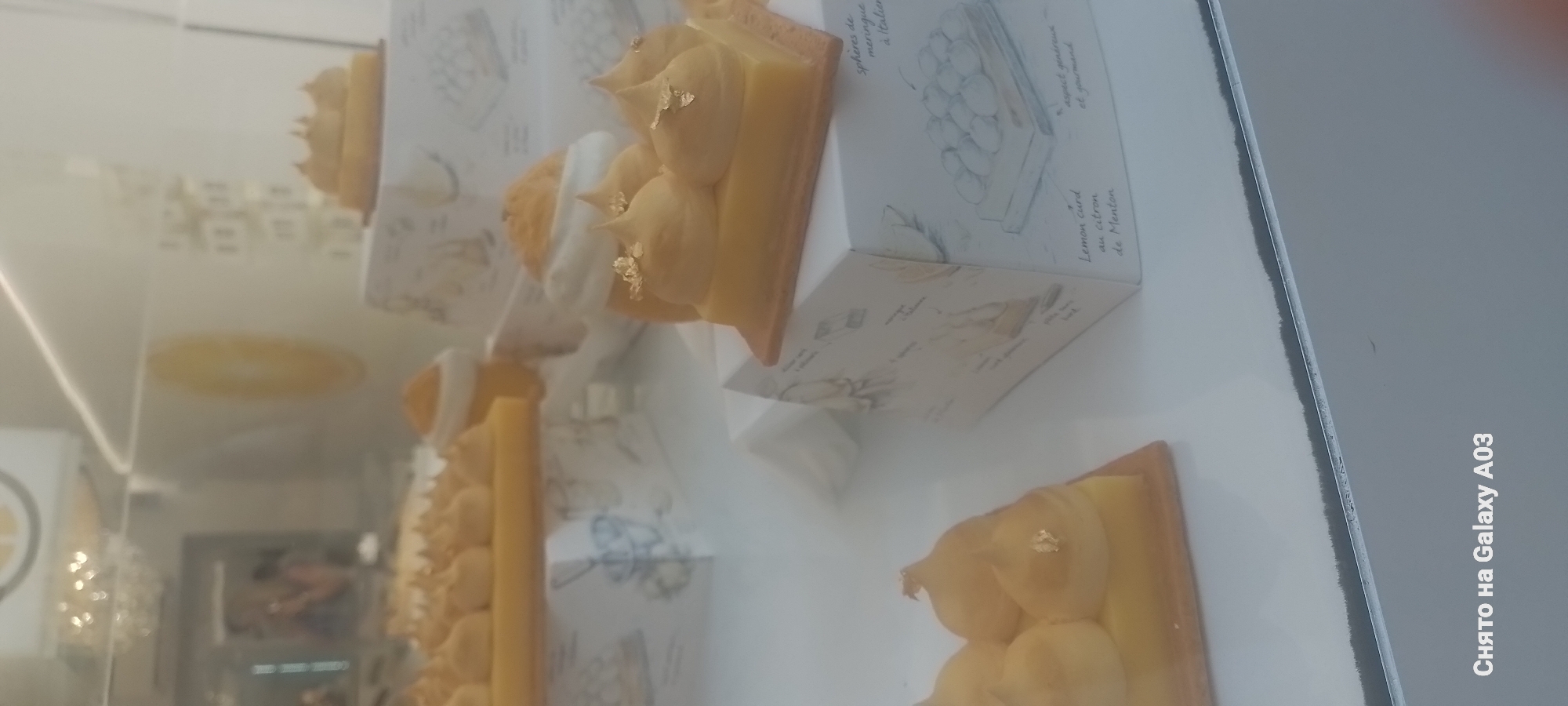 The image displays a close-up view of several lemon-themed desserts presented in what appears to be a bakery or pastry shop display case. The desserts are a vibrant yellow, some topped with meringue and flecks of edible gold leaf. One dessert is a rectangular tart with piped meringue peaks, while another is a small square pastry. The desserts are placed on decorative white boxes that feature line drawings of pastries and text, including "spheres de meringue à l'italienne," "Lemon curd au citron de Menton," and "aspect généreux et gourmand." The background is blurred, showing reflections and hints of lighting fixtures, suggesting an indoor setting. The text at the bottom of the image, "Снято на Galaxy A03," indicates the photo was taken with a Samsung Galaxy A03 phone. Although the text on the box mentions "Menton," the image itself does not provide specific visual cues to pinpoint the exact location within the city of Menton, France; it only suggests the desserts are inspired by or from Menton. There are no people clearly visible or identifiable in the frame.