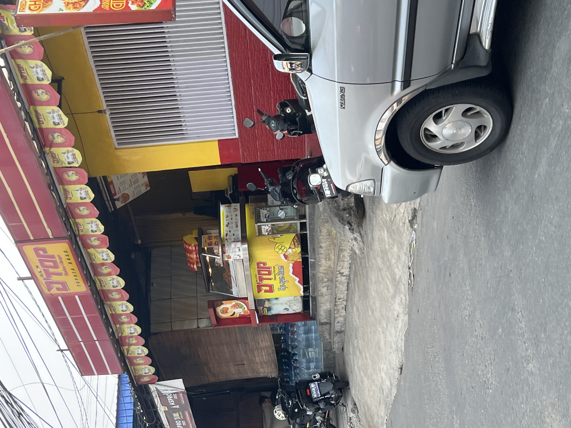 The image captures a street scene in Citeureup, Indonesia, featuring a food stall with a prominent red and yellow awning. The stall displays various food items and menus, with a "D'TOP Chicken & Burger" sign visible. Two motorcycles are parked in front of the stall, and a silver car is partially visible in the foreground, obscuring the lower right portion of the scene. The area appears to be a commercial or roadside setting, with a sidewalk and a paved street.