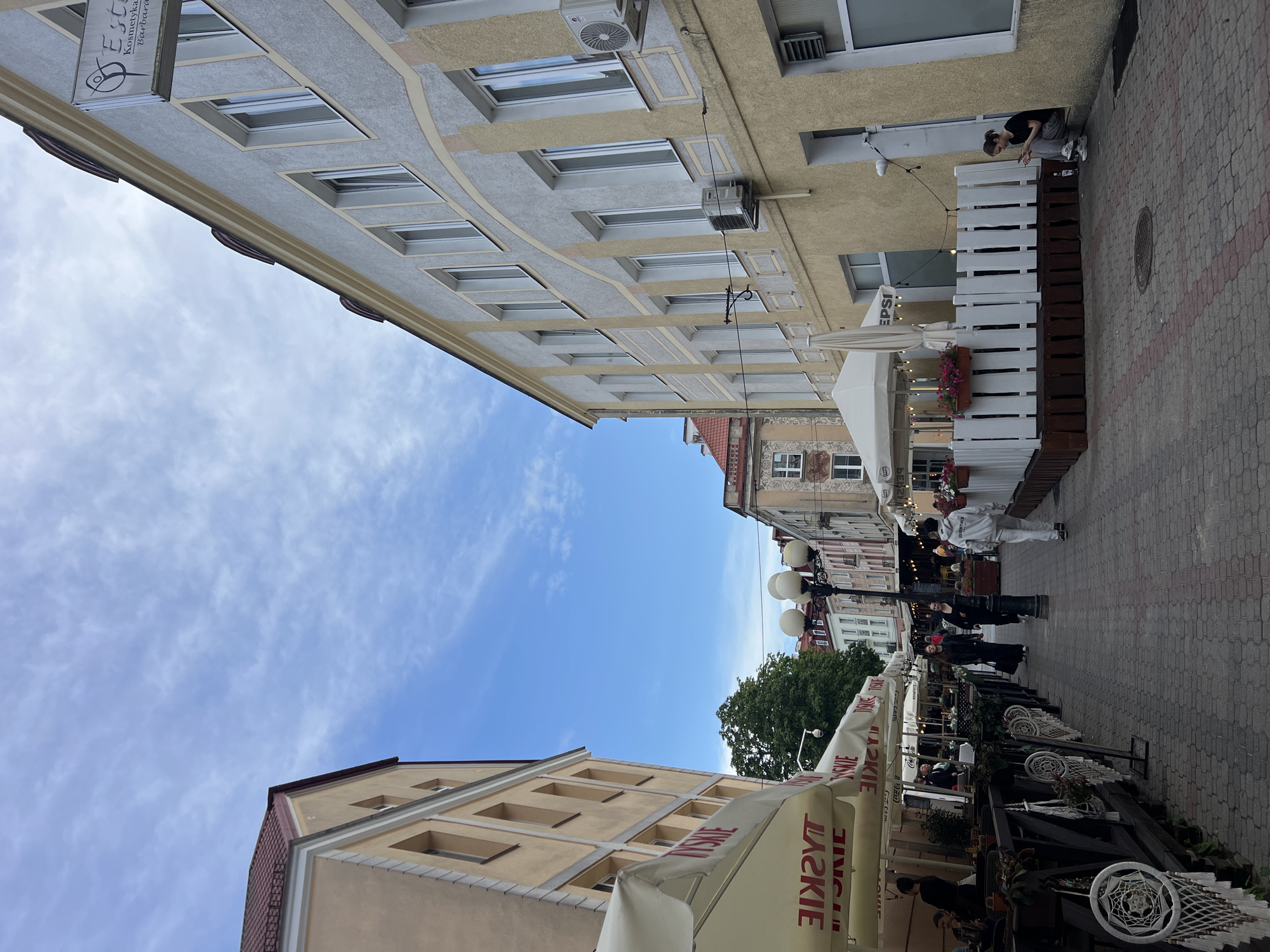The image depicts a street scene in Białystok, Poland, featuring outdoor seating areas of cafes or restaurants. On the left, a building with a light yellow facade has awnings with the "TYSKIE" logo. Tables and chairs are set up under these awnings, and decorative dreamcatchers hang from a railing. Further down the street, a white gazebo with the "PEPSI" logo is visible, also with outdoor seating enclosed by a white picket fence and planters with pink flowers.
On the right, a building with a pale yellow and grey facade has multiple windows. An air conditioning unit is mounted on the exterior wall. A young woman is seated on the ground near a doorway, looking at her phone, with a bottle and a glass on the ground beside her.
Several people are walking along the cobblestone street. The sky above is blue with scattered white clouds. The street is lined with buildings and lampposts, indicating a city environment.