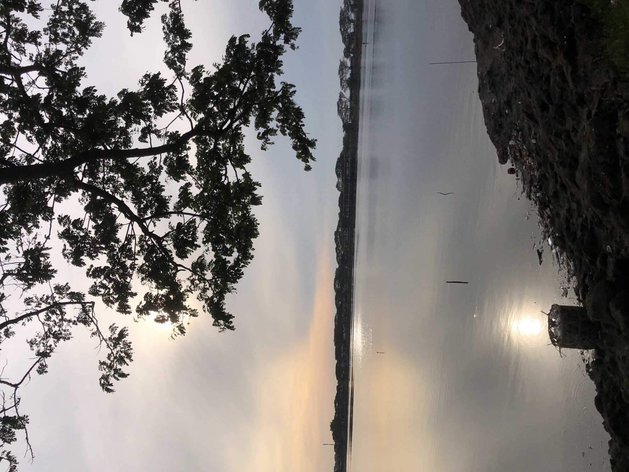 A wide shot captures a serene lake at dusk, with a silhouetted tree canopy framing the upper portion of the image. The sun, partially obscured by clouds, casts a gentle glow across the water, creating a shimmering reflection. In the background, a line of buildings and trees stretches along the opposite shore, suggesting a suburban or semi-rural setting. The foreground is a rough, debris-strewn embankment, with a woven basket partially filled with trash resting near the water's edge. The overall atmosphere is tranquil, with a hint of environmental neglect due to the litter. The location is likely the Lhokseumawe Reservoir in Lhokseumawe, Indonesia, known for its scenic beauty and recreational opportunities.