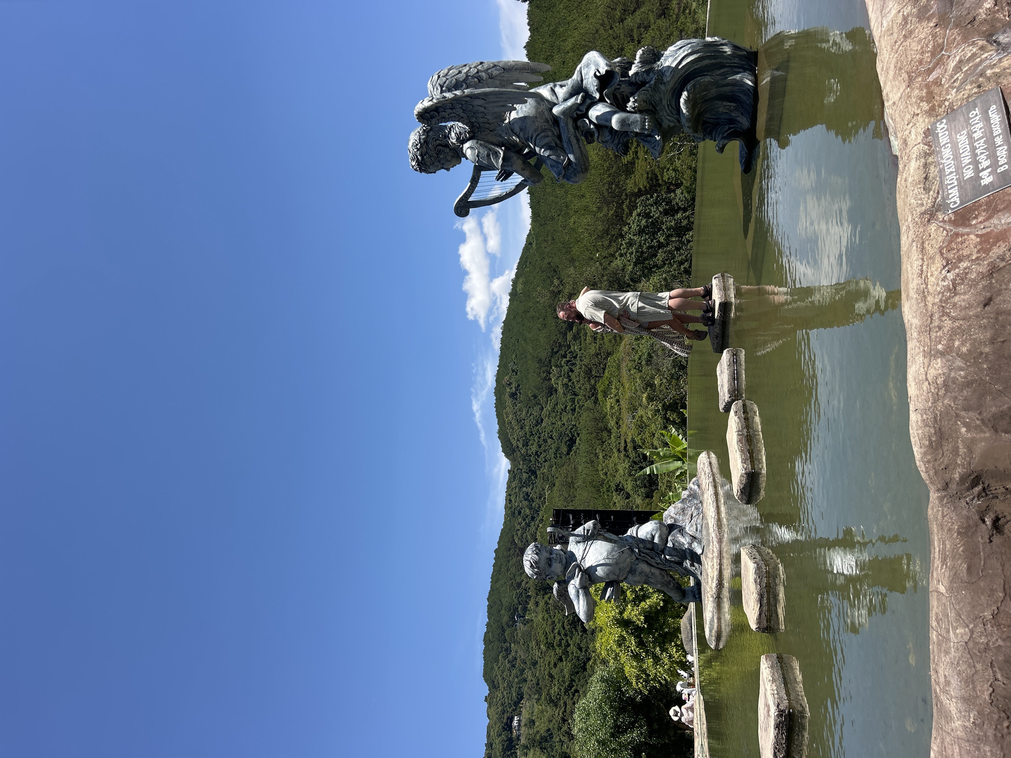 A bright, sunny day in Da Lat, Vietnam, captures a picturesque outdoor scene featuring a tranquil pond, ornate statues, and lush green hills. In the mid-ground, a person, dressed in light-colored clothing, stands on rectangular stepping stones that cross a body of water, observing the surroundings.
To the left of the pond, a large, dark-colored statue of an angel playing a harp stands prominently. Further to the right, another statue, resembling a child or Cupid with a bow and arrow, is partially visible near the water's edge. These intricate statues appear to be made of metal or stone, lending a classical aesthetic to the natural setting.
The background is dominated by rolling, densely forested hills under a clear, vibrant blue sky, dotted with a few fluffy white clouds. The water in the pond reflects the sky and parts of the surrounding greenery. Along the right edge of the pond, a textured stone wall or embankment is visible.
A black rectangular sign affixed to this stone wall displays a warning in multiple languages: "CÁM LỘI XUỐNG NƯỚC" (Vietnamese), "NO WADING" (English), "물에 들어가지 마십시오" (Korean), and "В воду не входить" (Russian), clearly indicating that entering the water is prohibited. The scene depicts a peaceful moment, likely in a garden or park setting, during what appears to be midday given the strong sunlight and clear skies.