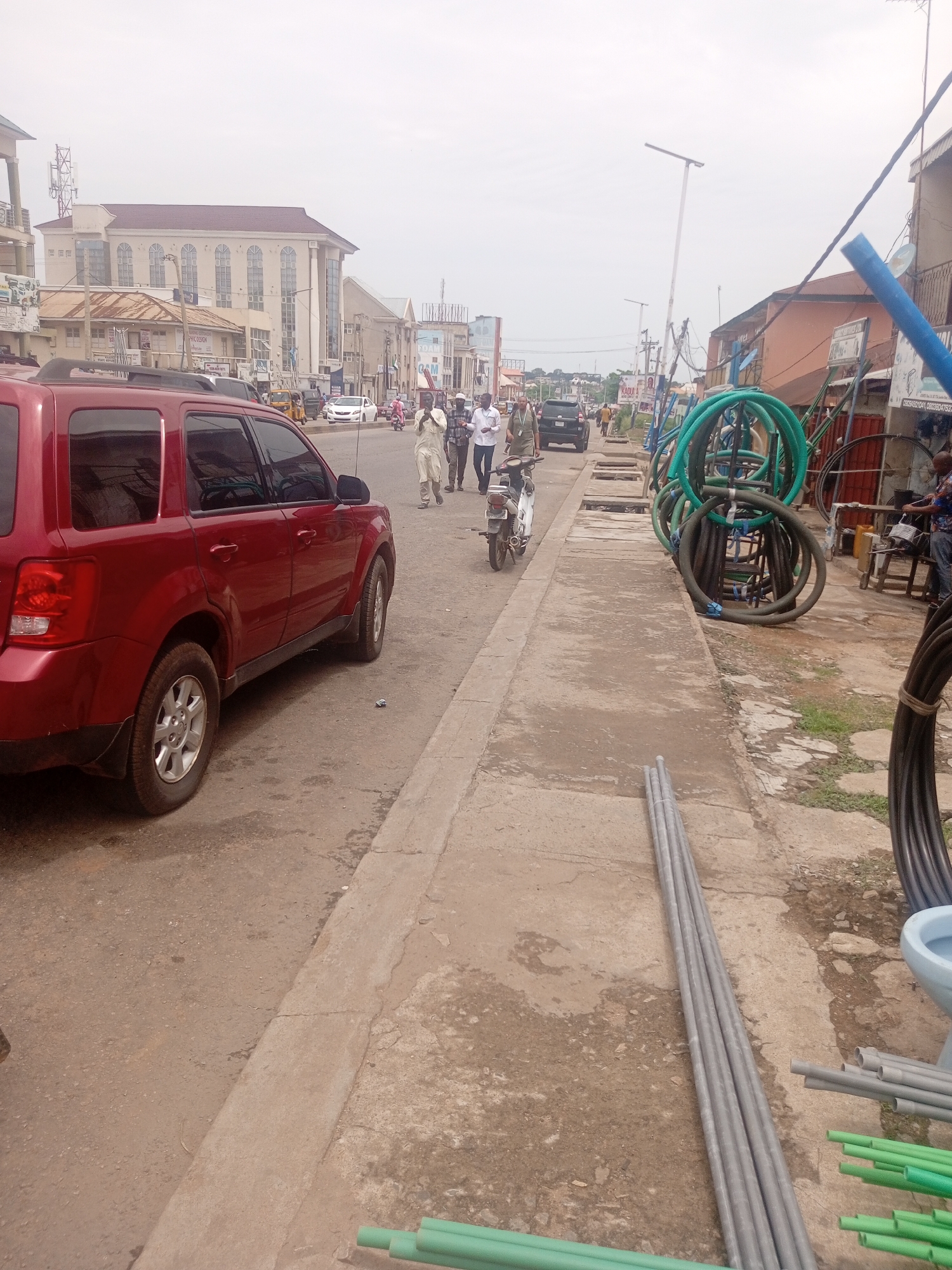 In the city of Dadi Riba, Nigeria, a roadside scene unfolds under an overcast sky. A red SUV is parked on the left side of a paved road, its passenger side facing the viewer. A concrete sidewalk runs parallel to the road, separating it from a collection of materials. On the sidewalk, bundles of green and gray pipes are stacked, alongside coiled green hoses and what appears to be a light blue toilet bowl. Further down the sidewalk, a person is partially visible in the background, seemingly working at a stall or shop.
Across the road, several individuals are walking, with one person on a motorcycle moving away from the camera. In the distance, buildings line the street, including a large, light-colored structure with arched windows and a prominent antenna tower. Traffic, including cars and a tricycle, is visible on the road, suggesting active movement within the urban environment. The overall impression is one of a bustling commercial street with construction or plumbing supplies being displayed for sale.