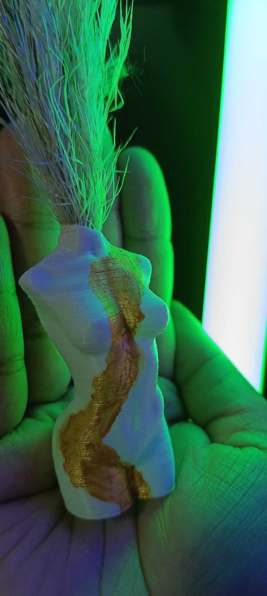 A 3D-printed statuette of a female torso, colored white with gold accents, is held in a person's hand. The statuette has a texture indicating it was 3D printed in layers. Green and blue light illuminates the scene, creating a dramatic effect. The background is dark, with a bright white vertical light source on the right. There is no indication of Wels, Austria.