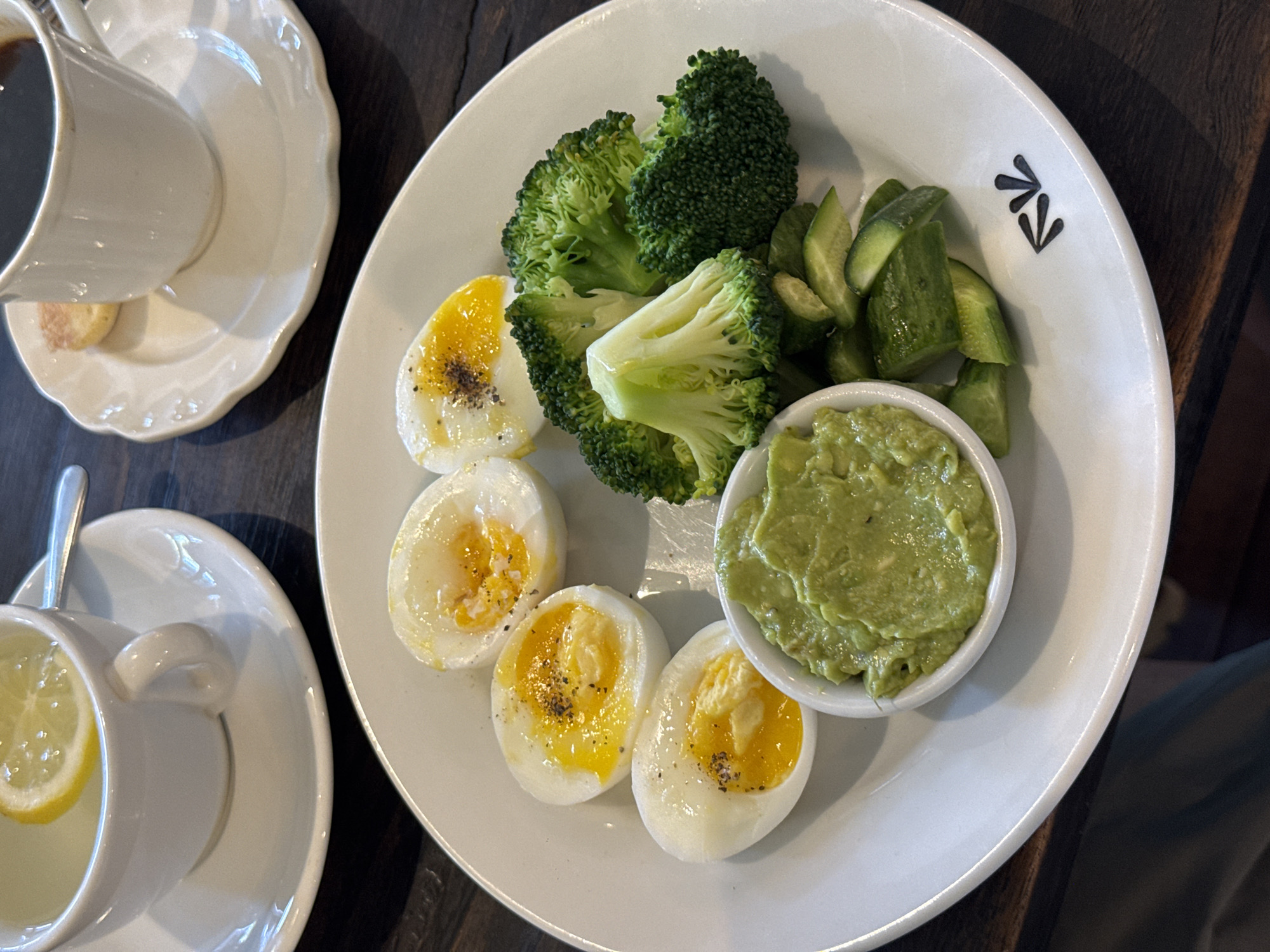 This high-angle, close-up photograph captures a well-balanced meal laid out on a dark wooden table, characteristic of a cafe or restaurant in North Kuta, Indonesia.
The central subject is a large white oval plate filled with wholesome food items. Six halves of hard-boiled eggs are arranged on the plate, their bright yellow yolks still slightly soft and seasoned with freshly ground black pepper. Alongside the eggs are several large florets of steamed or blanched broccoli, showcasing their vibrant green color. A portion of sliced green vegetables, appearing to be cucumbers, is also present. A small white ceramic bowl on the plate holds a creamy green dip, consistent with guacamole or mashed avocado. A subtle black graphic logo, resembling a series of stylized arrows or leaves, is visible on the rim of the main plate.
Accompanying the meal are two white ceramic cup-and-saucer sets. In the upper left of the frame, a cup contains a dark liquid, likely coffee, with a small biscuit or cookie resting on its saucer. In the lower left, another cup holds a light yellow beverage, identifiable as lemon tea, complete with a lemon slice and a spoon.
The scene is brightly lit, suggesting it is daytime. The overall impression is one of a healthy and carefully prepared breakfast or brunch. No people are visible in the image.
