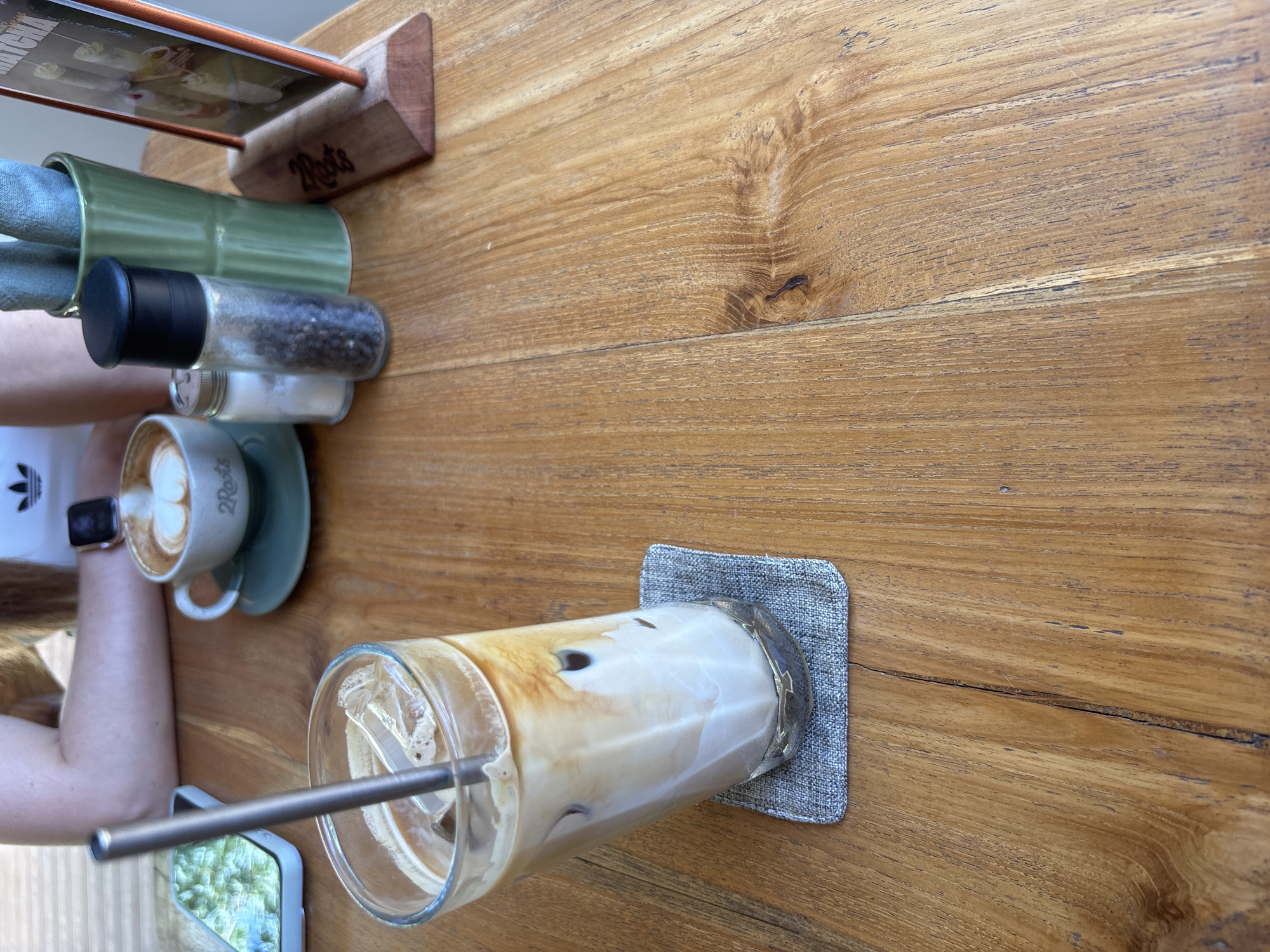 The scene is captured on a wooden table, likely in Pecatu, Indonesia, given the presence of "2 Roots" branding, which is associated with a café in that area. The table features drinks: a glass of iced coffee with a metal straw, and a cup of latte with milk art on a green saucer. The coffee is accompanied by condiments such as salt and pepper shakers, and a napkin holder. There is also a phone. An arm is visible, clad in a white Adidas t-shirt. The composition is shot from an overhead angle.