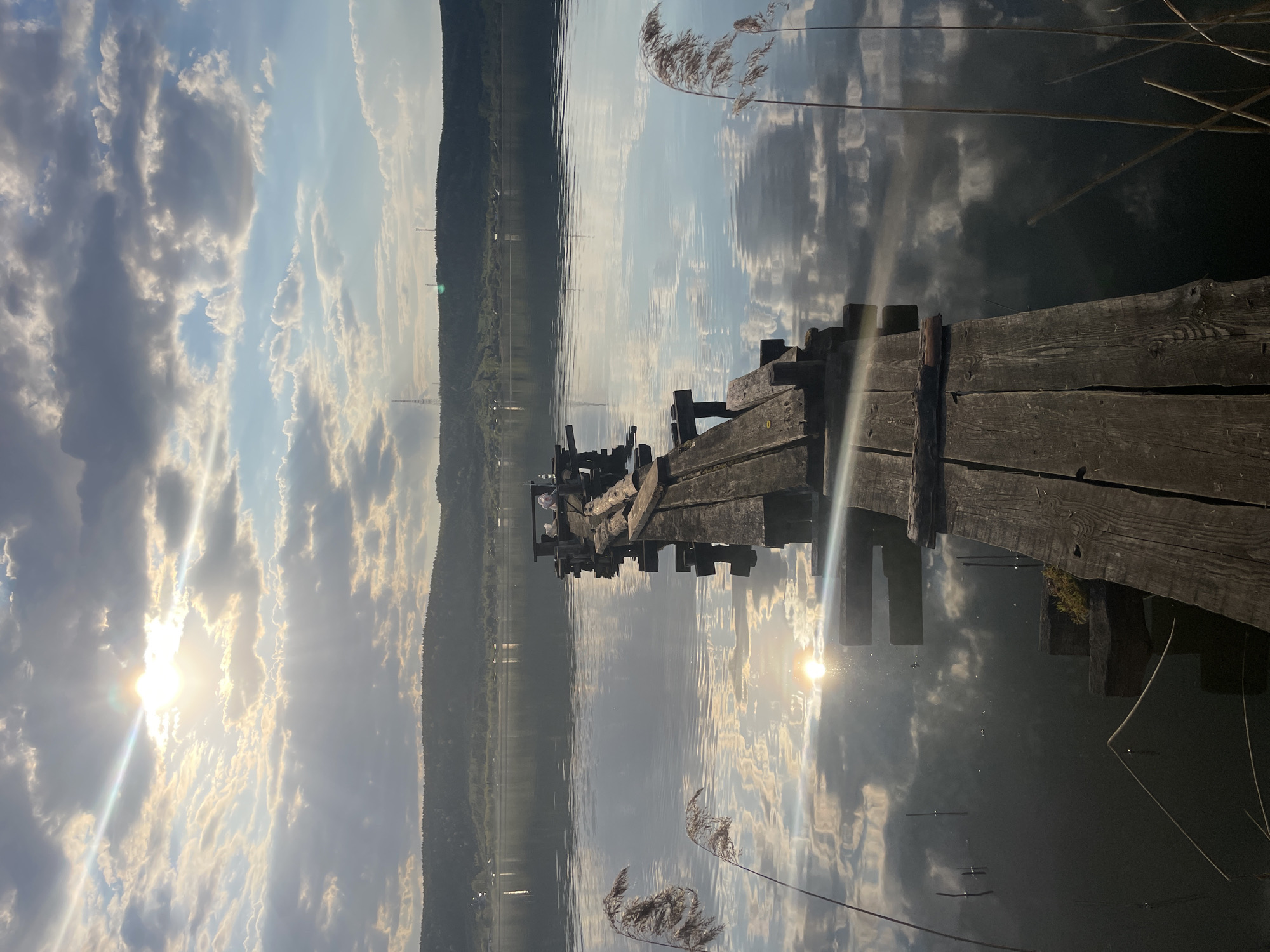 The image shows an old, weathered wooden pier extending into a calm body of water. The pier appears to be in a state of disrepair, with missing planks and supports. The sun is low in the sky, reflecting off the water and creating a glare. The sky is filled with clouds, which are also reflected in the water. Reeds are visible in the foreground. In the distance, a forested hillside can be seen. The image is likely taken at Suksun, Russia, based on the prompt.