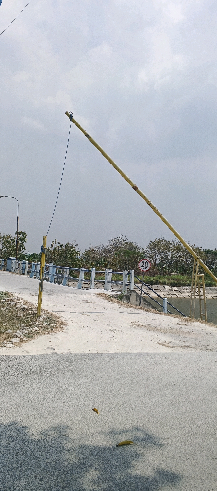 The image shows a barrier gate, a bridge, and a canal in Parengan, Indonesia. The barrier gate is yellow and extends across a road. The bridge is made of concrete and has a railing. The canal is filled with water and has vegetation on its banks. There are no people visible in the image. The sky is cloudy, and the sun is shining.