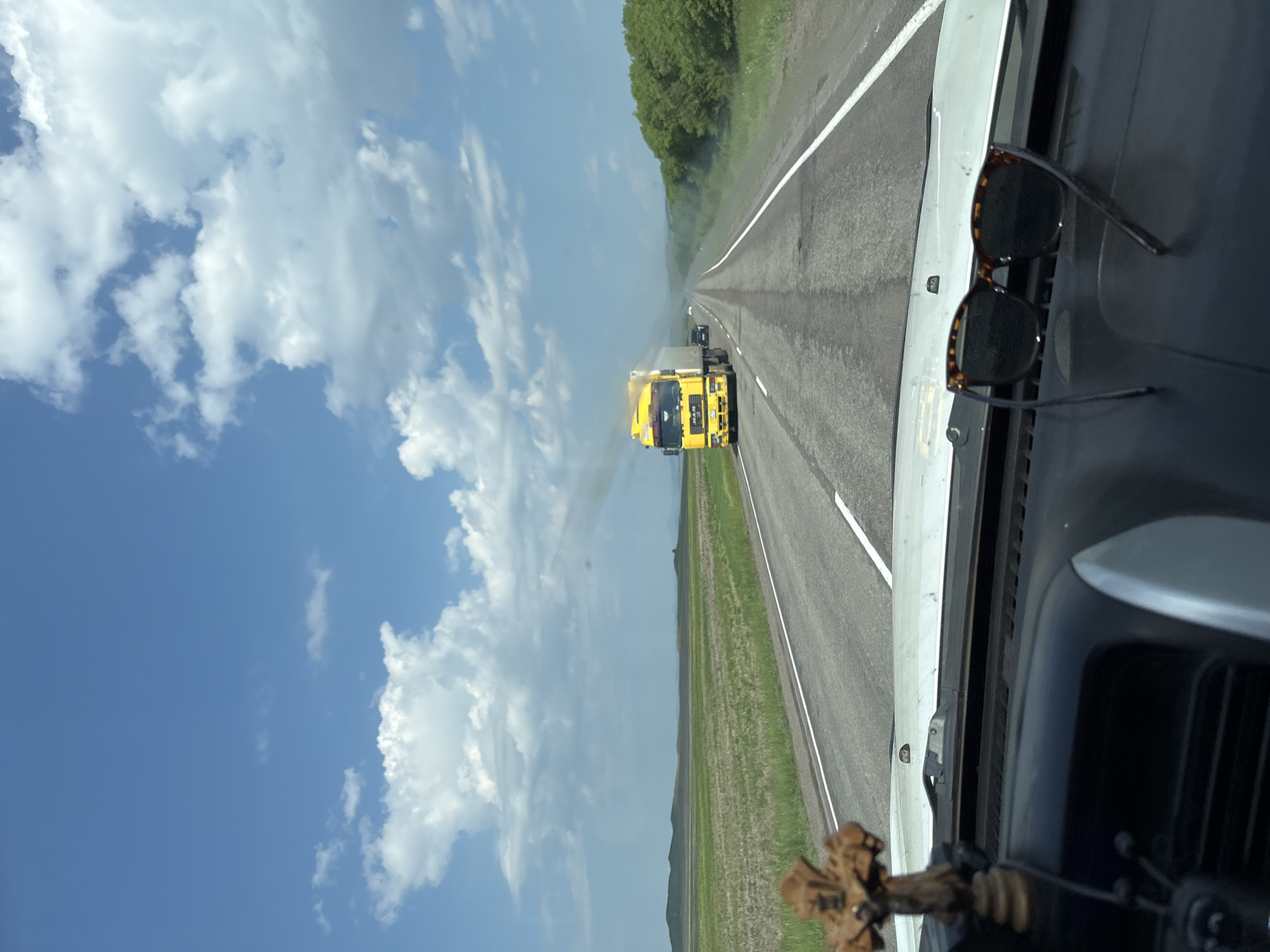 The image is a view from inside a vehicle, looking out at a rural highway. The road is asphalt with white lane markings, and it stretches into the distance. On the left side of the road is a green field with some dry grass. On the right side of the road is a dense line of green trees. In the distance, a yellow truck with "MAN" written on it is driving on the highway, emitting dark smoke from its exhaust. There are other vehicles on the road ahead, and the sky is blue with scattered white clouds. In the foreground, on the dashboard of the vehicle, are a wooden crucifix and a pair of sunglasses. The scene appears to be on a sunny day in a rural area. The location is identified as the city of Borodino, Russia.