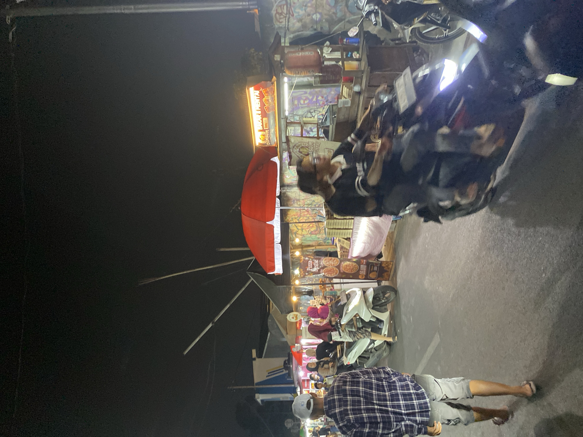 This is a night scene at a street food market in the city of South Tangerang, Indonesia. A man in a plaid shirt and shorts walks away from the camera on the left side of the image. In the background, several food stalls are set up with awnings and bright lighting. One stall has a red umbrella and displays pizzas on a menu. Another stall has a lit sign that reads "BAKAR LENTAI" and features pictures of grilled food. Two people are riding a motorcycle in the center of the frame, moving from right to left. Several other people are visible near the food stalls, some wearing headscarves. Various objects such as coolers, umbrellas, and stacks of items are present at the stalls. The ground is paved asphalt.