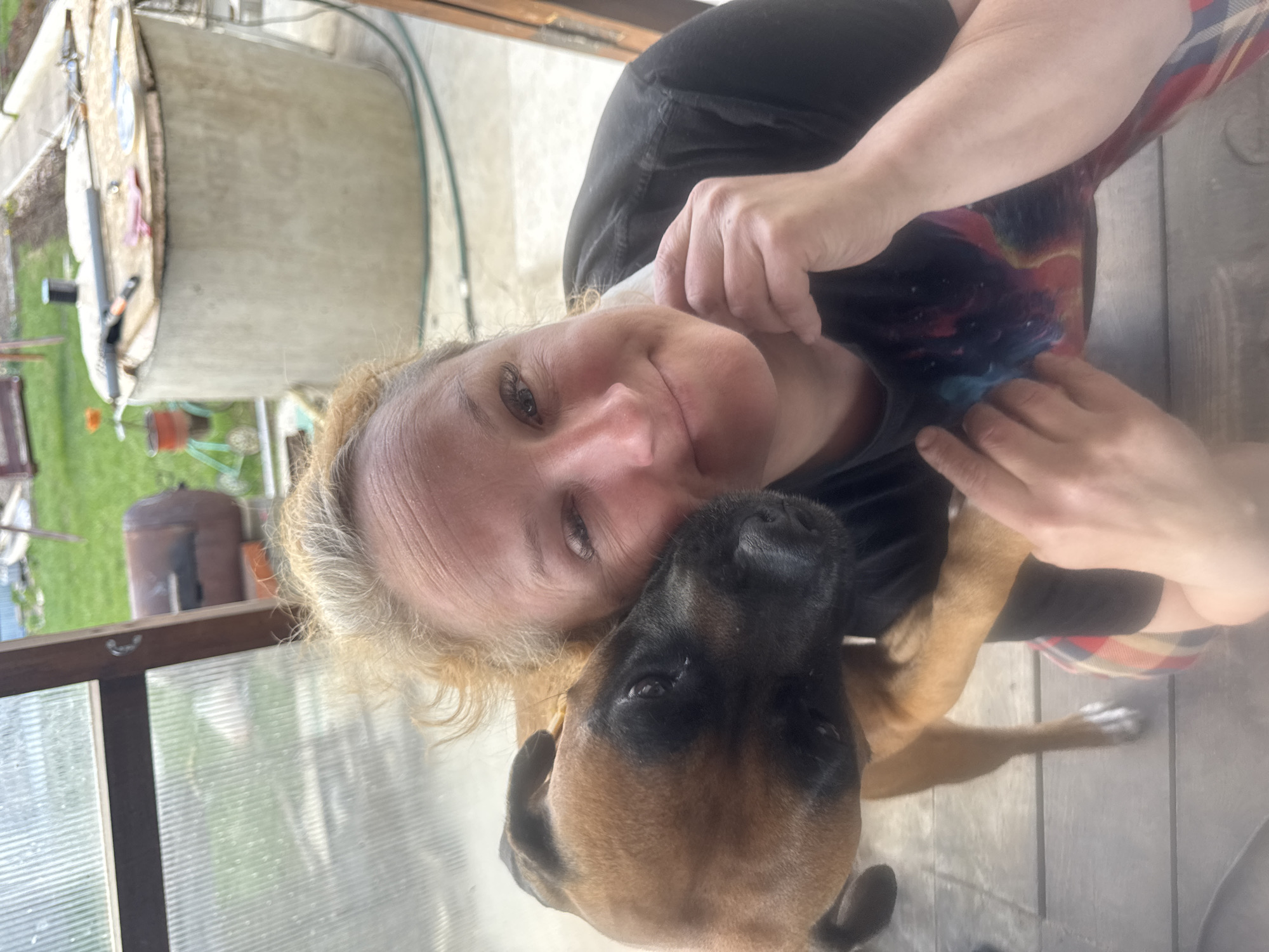 A woman is pictured in close proximity to a brown boxer dog, likely in an embrace. The woman has fair skin and blonde hair; she is wearing a black top. The dog's head is nestled against the woman's face. The setting appears to be outdoors, with a blurred background of what could be a garden or yard. The location is said to be in the city of Chaikovsky, Russia. The exact setting in the city is not visually verifiable. There is a greenhouse-like structure visible on the left side of the frame.