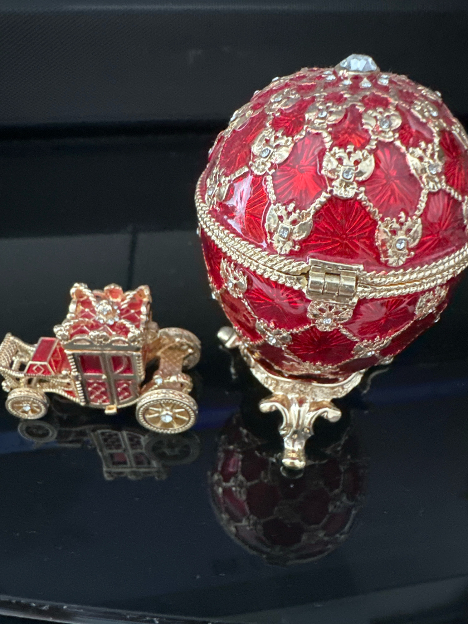The image shows two decorative objects, a red and gold ornate egg and a smaller red and gold carriage, placed on a reflective dark surface. The egg is on a stand and appears to be a music box or a trinket box, adorned with intricate gold filigree and small clear stones. The carriage is to the left of the egg, also detailed with gold trim and red accents. Both objects are highly decorative and appear to be made of metal, possibly with enamel or painted surfaces. The background is dark and out of focus, suggesting a controlled lighting environment. There is no indication of the location being in the city of Minsk, Belarus.