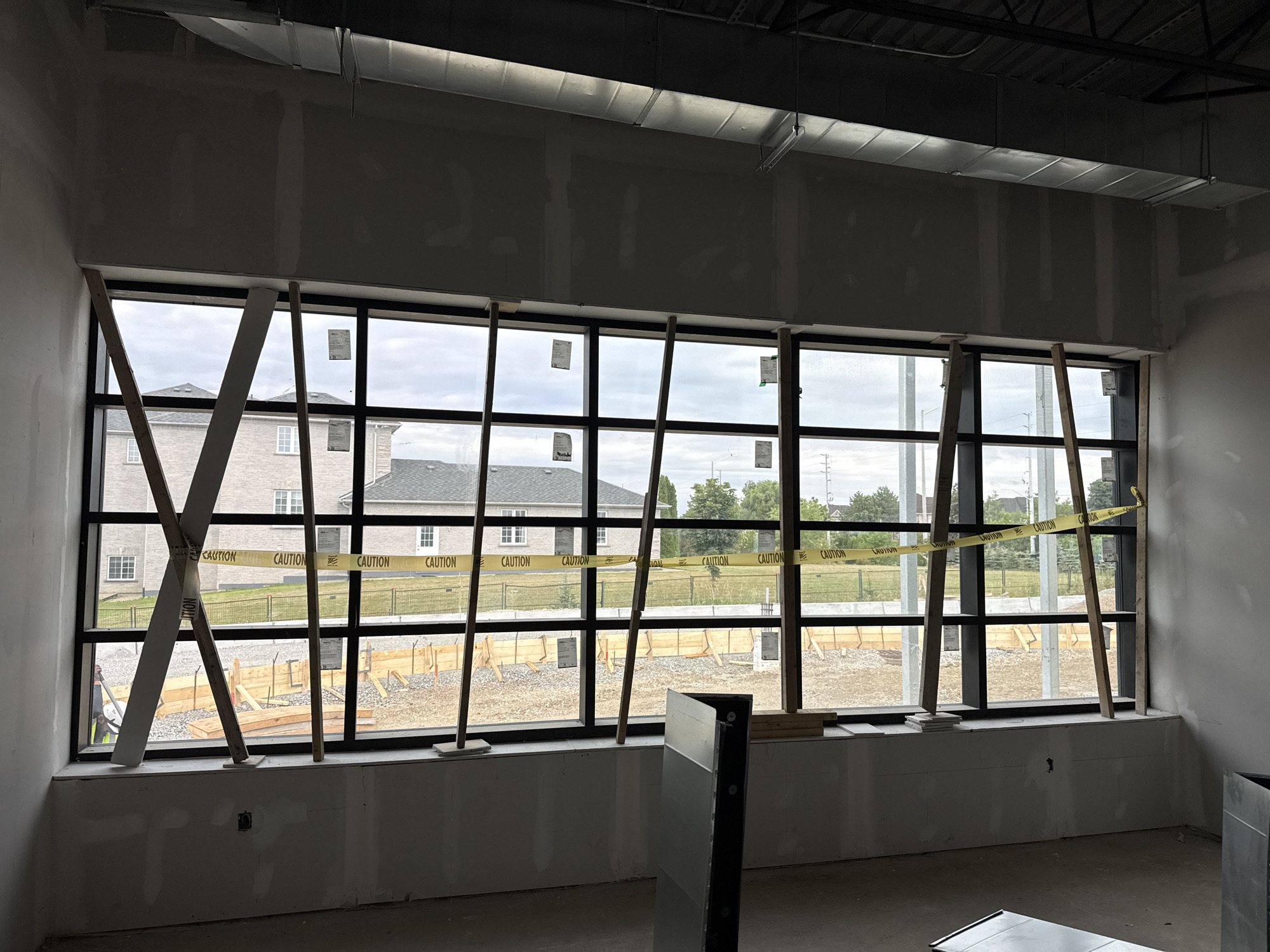 The image shows the interior of a building under construction in Brampton, Canada. A large window runs along the wall, offering a view of a residential neighborhood with brick houses and trees. Yellow caution tape is stretched across the window, with "CAUTION" printed repeatedly. Several wooden planks are propped against the window from the inside, some crossing each other. In the foreground, a partially visible metal object stands upright. The walls appear to be finished with drywall, and a large ventilation duct is visible on the ceiling. The floor is bare concrete.