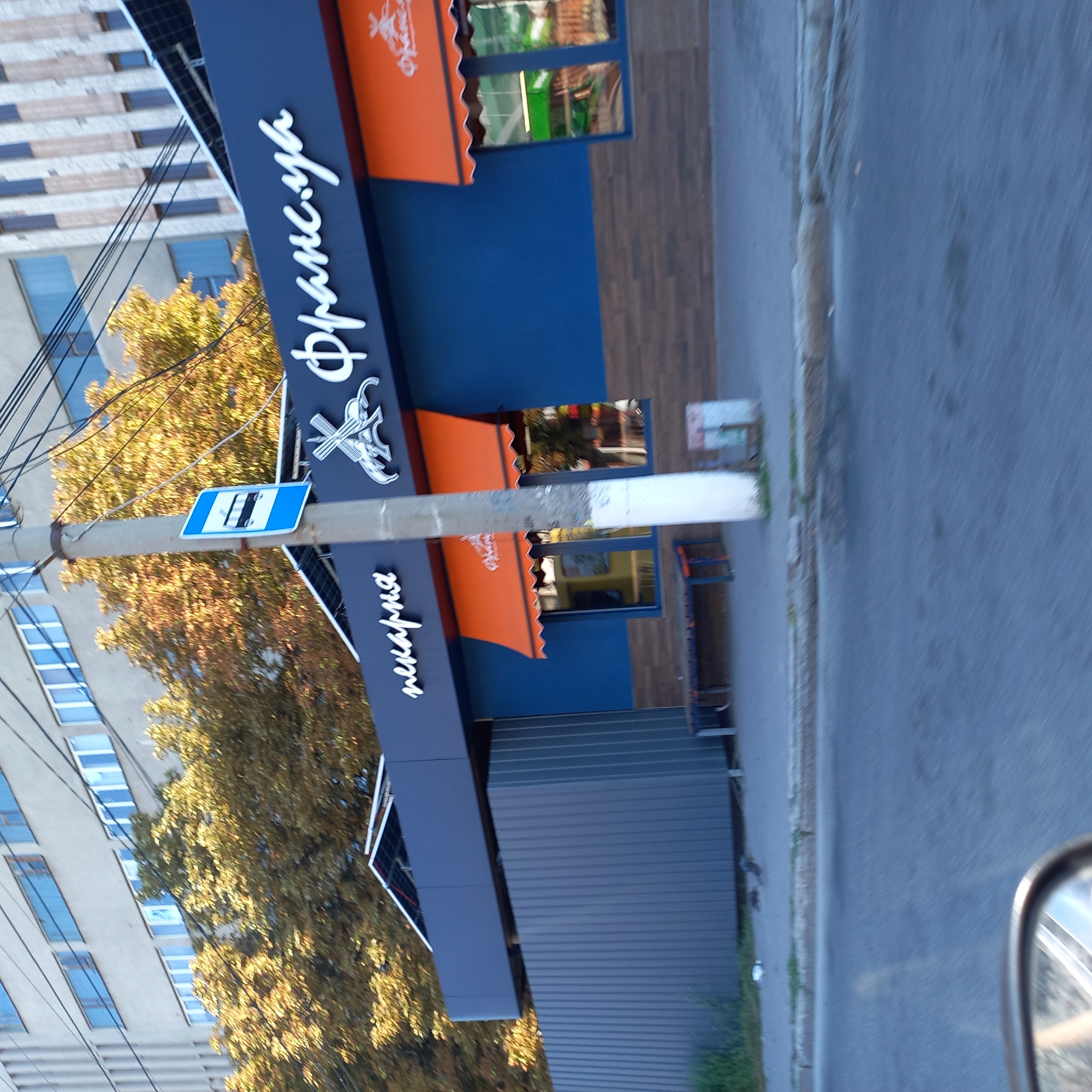 The image shows the exterior of a bakery named "Франс.уа" located in Kropyvnytskyi, Ukraine. The bakery has a blue facade with orange awnings over the windows. The name of the bakery is displayed in white, stylized script on the dark blue upper part of the building. A bus stop sign is visible on a pole in front of the bakery. Behind the bakery, trees with autumn foliage are visible, and a multi-story building stands in the background. A metal fence runs along the side of the building. The scene is captured from a low angle, likely from a moving vehicle, as indicated by the blurred foreground and reflection in a rearview mirror on the bottom left. There are no visible people in the image.