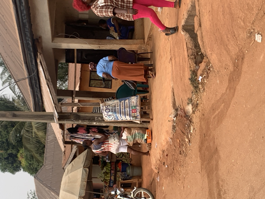 The image depicts an outdoor commercial scene in Otuocha, Nigeria. The foreground features an unpaved reddish-brown dirt road, uneven with depressions and a shallow drainage channel containing debris on the right. In the midground, a wooden utility pole stands centrally with power lines extending across the frame.
On the left, a market stall operates under a light-colored umbrella canopy, displaying fruits and vegetables in baskets on a wooden stand. A silver motorcycle's front wheel is visible on the far left. Adjacent to the market stall, a woman with dark hair, wearing a white top patterned with red and blue (resembling the US flag) and white shorts, stands barefoot, her left arm raised. A large woven sack and an orange crate rest near her feet. Behind her, a rack displays various clothing items.
Centered in the midground is a blue and white "CYNTHIA POS" sign, advertising financial services such as withdrawals, transfers, deposits, and data/airtime sales, with a phone number (09136198207) displayed. The sign is propped on a wooden easel.
To the right of the sign, a light-colored building with an open doorway reveals two individuals seated inside. Outside this building, a woman wearing a light blue top and a long reddish-brown skirt walks away from the camera, towards the building's entrance. Several plastic chairs, including green and purple ones, are positioned near the building's entrance. Further right, a woman with bright red curly hair, wearing a plaid shirt and bright pink leggings with black slides, walks from right to left, partially obscured.
The background shows multiple structures with corrugated metal roofs, some rusted brown and others grey, along with tropical foliage and palm trees under an overcast sky.