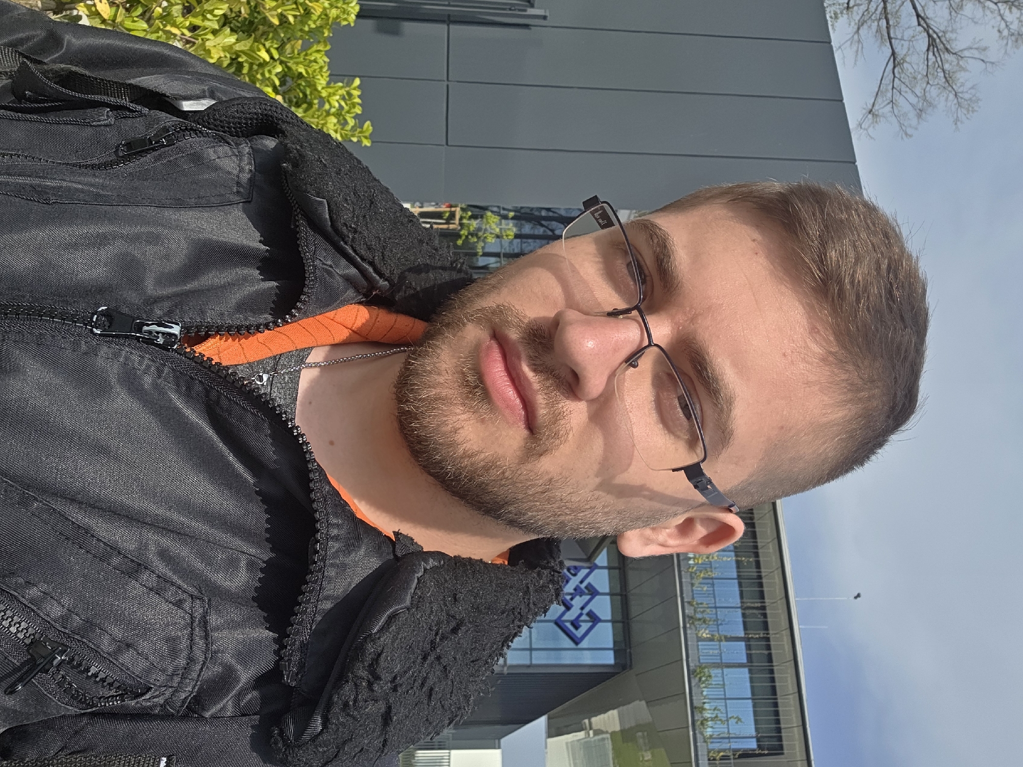 A fair-skinned man with short brown hair and a beard is the focus of the image, wearing glasses and a black jacket with an orange lining. He stands outdoors, likely in front of the UniCredit Bulbank building, a financial institution, identifiable by its logo in the background, which places the location in Plovdiv, Bulgaria. The time of day appears to be daylight, given the light and shadows. Some greenery is visible in the upper left corner, suggesting a park or planted area nearby. The background includes a modern building with glass windows and a grey facade.