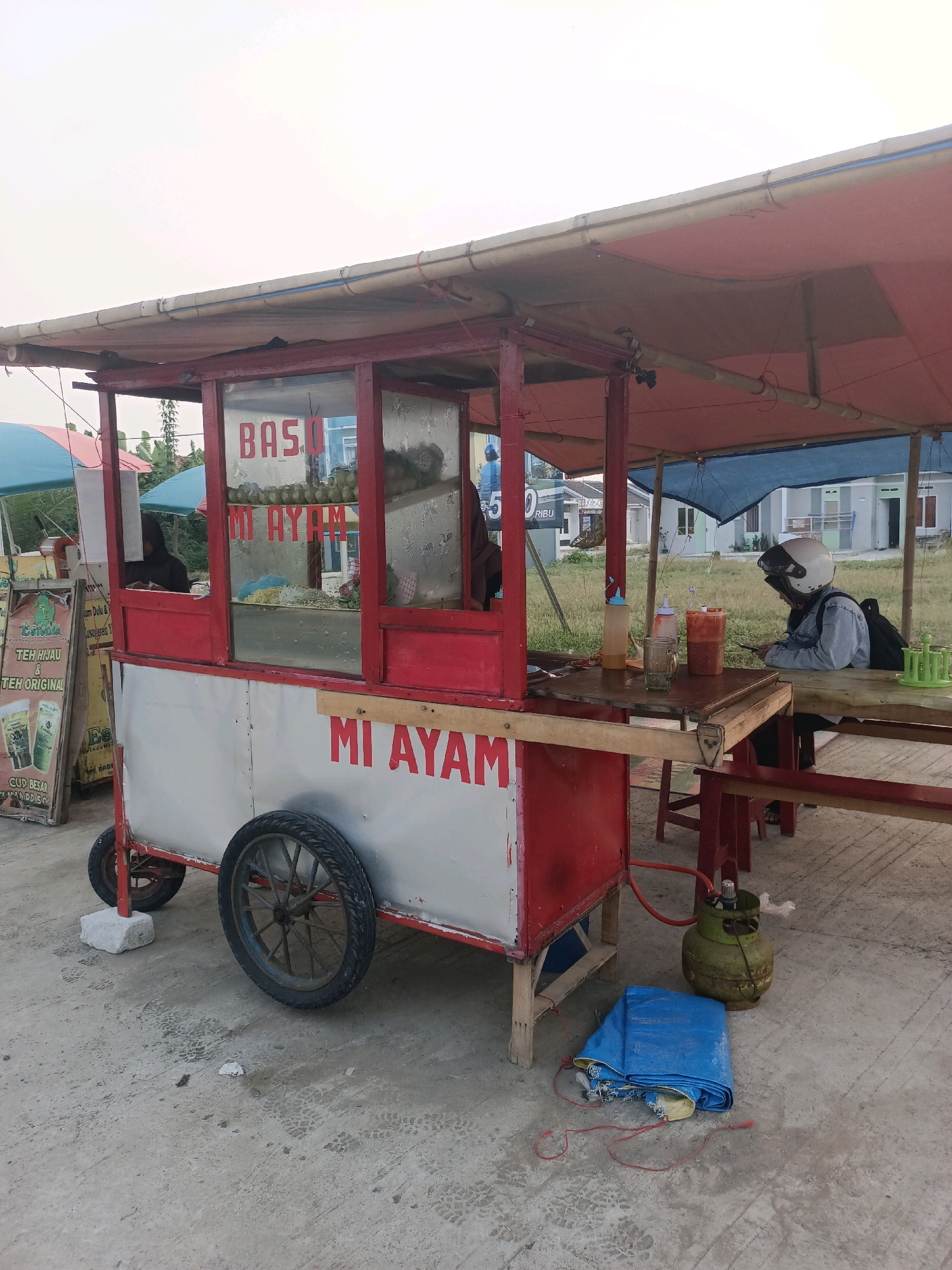 The image depicts an outdoor food stall, likely a mobile cart, set up on a paved surface. The stall itself is primarily red and silver, with glass panels showcasing food items. Prominently displayed on the stall are the words "BASO" and "MI AYAM," indicating the food being sold. A blue tarp and a green gas cylinder are visible on the ground near the stall.
To the left of the main stall, another food stall is visible with a sign advertising "TEH HIJAU & TEH ORIGINAL" and "CUP BESAR." Two people are partially visible within the main food stall, appearing to be working.
To the right of the food stall, a long wooden table is set up with chairs. A person wearing a helmet and a denim jacket is seated at the table, looking at a mobile phone. There are condiments and drinks on the table, as well as a rack of green glasses.
In the background, several houses with light-colored facades and red roofs are visible, suggesting a residential area. The sky is overcast, indicating potentially hazy or cloudy weather. The overall environment suggests a casual, roadside food vendor setup in a semi-urban or suburban setting in Karawang, Indonesia. The presence of houses and the paved ground indicate it's likely within a populated area.