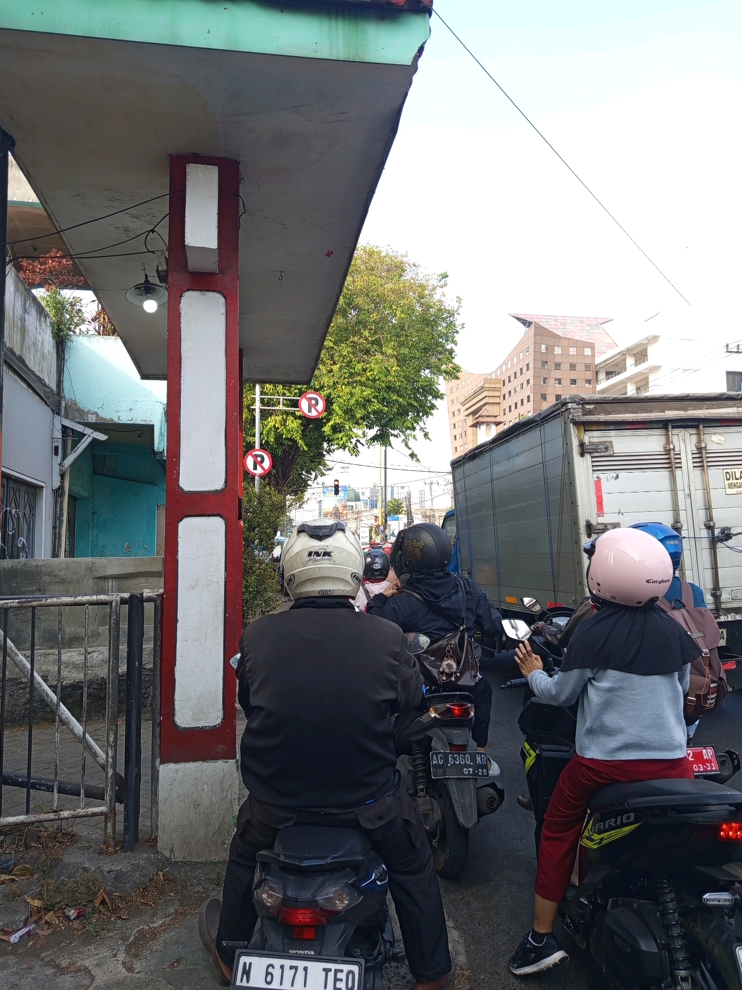 The image depicts a traffic jam in the city of Malang, Indonesia. Several motorcycles are visible in the foreground, with riders wearing helmets. One rider wears a white helmet, another a black helmet, and a third a pink helmet. The motorcycles are stopped in traffic, with a large truck and other vehicles further ahead. In the background, there are buildings and trees. Two "no parking" signs with red circles and a diagonal line over the letter "R" are mounted on a pole. A lamppost with a single bulb hangs from an overhead structure. The scene appears to be on a street in a developed urban area.