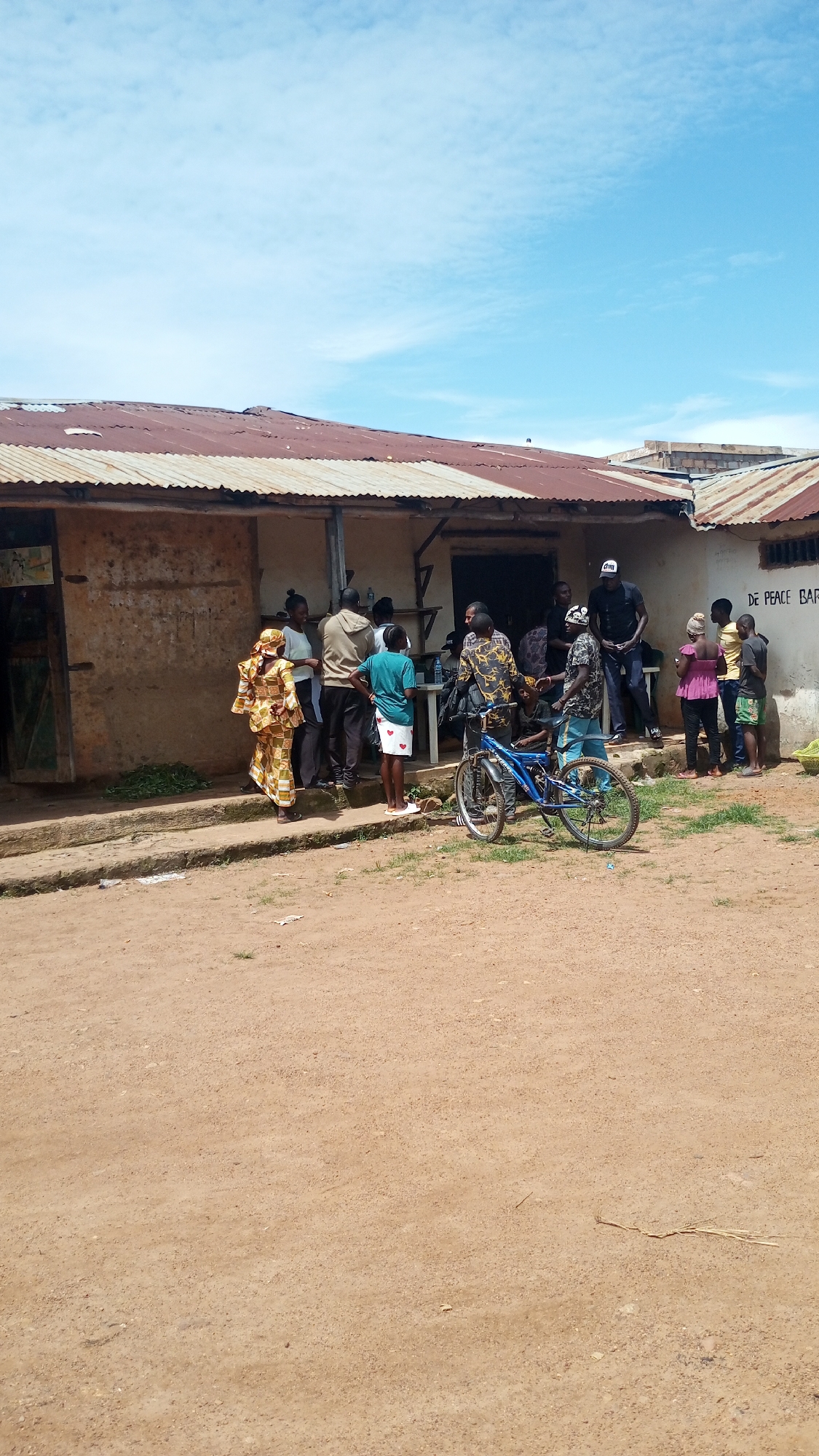 The image captures an outdoor scene in Vom, Nigeria, likely in front of a local business. A group of approximately ten individuals, of varying ages and genders, are gathered near a weathered, single-story building with a corrugated iron roof. Some people appear to be standing in a line or grouping in front of an open doorway or window of the building, possibly interacting with someone inside or observing a transaction.
On the right side of the image, a blue bicycle is propped up, leaning against the building or a person. The ground in front of the building is unpaved, composed of dirt and sand, with some scattered debris. Vegetation is visible along the base of the building. The sky above is blue with scattered white clouds. One of the buildings to the right has the text "DE PEACE BAR" painted on its exterior wall. The overall impression is of a casual, everyday gathering in a semi-urban or rural setting.