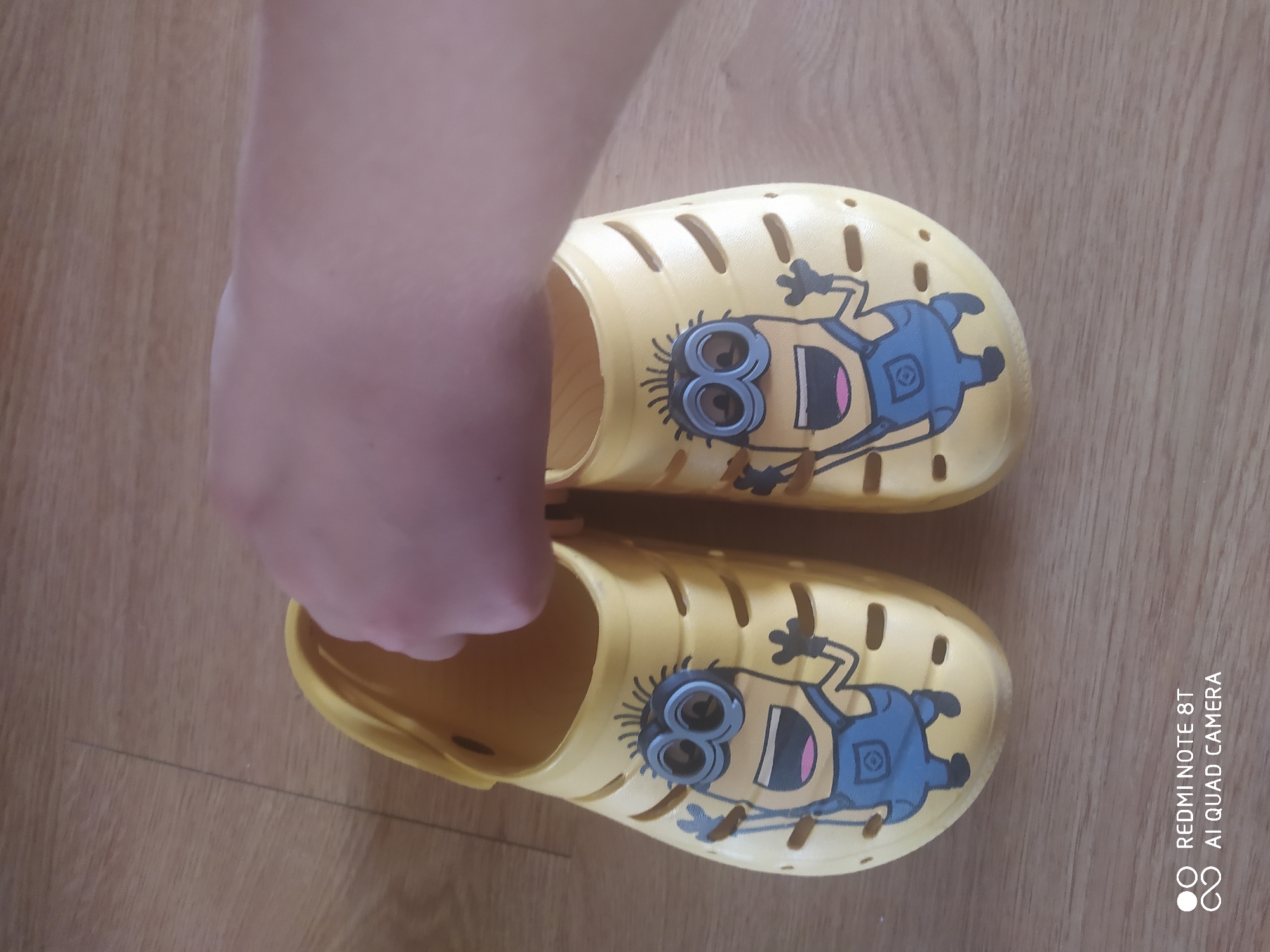 A pair of yellow child-sized clogs with a Minion character printed on each is shown. The clogs have an open heel and drainage holes along the top. A person's hand is seen holding the left clog from above, as if to put it on. The clogs are placed on a light brown wooden floor, which appears to be the floor of a room. The lighting suggests it could be daytime, indoors. There is no indication of weather or a specific location. The image includes a watermark in the bottom right corner which reads "REDMI NOTE 8T AI QUAD CAMERA".