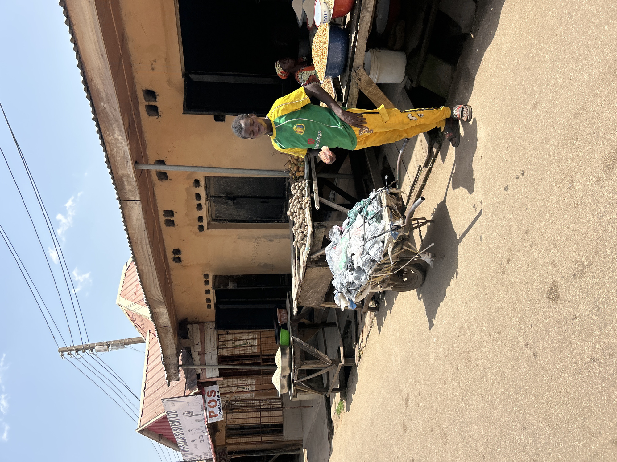 In the city of Jos, Nigeria, a young Black male stands next to a market stall, holding a piece of food in his right hand. He is wearing a green t-shirt with yellow accents and yellow pants. He is positioned in front of a stall with a yellow-orange facade, which has a corrugated metal roof. Behind him, a woman with a red headscarf is visible, seated behind a large blue bowl filled with yellow kernels. Adjacent to this stall, another stall displays mounds of potatoes on a wooden table. A two-wheeled cart, laden with a pile of bagged items, is situated in the foreground, to the left of the young man. The ground is paved with asphalt, and power lines are visible overhead against a bright blue sky. The scene appears to be an outdoor market on a sunny day.