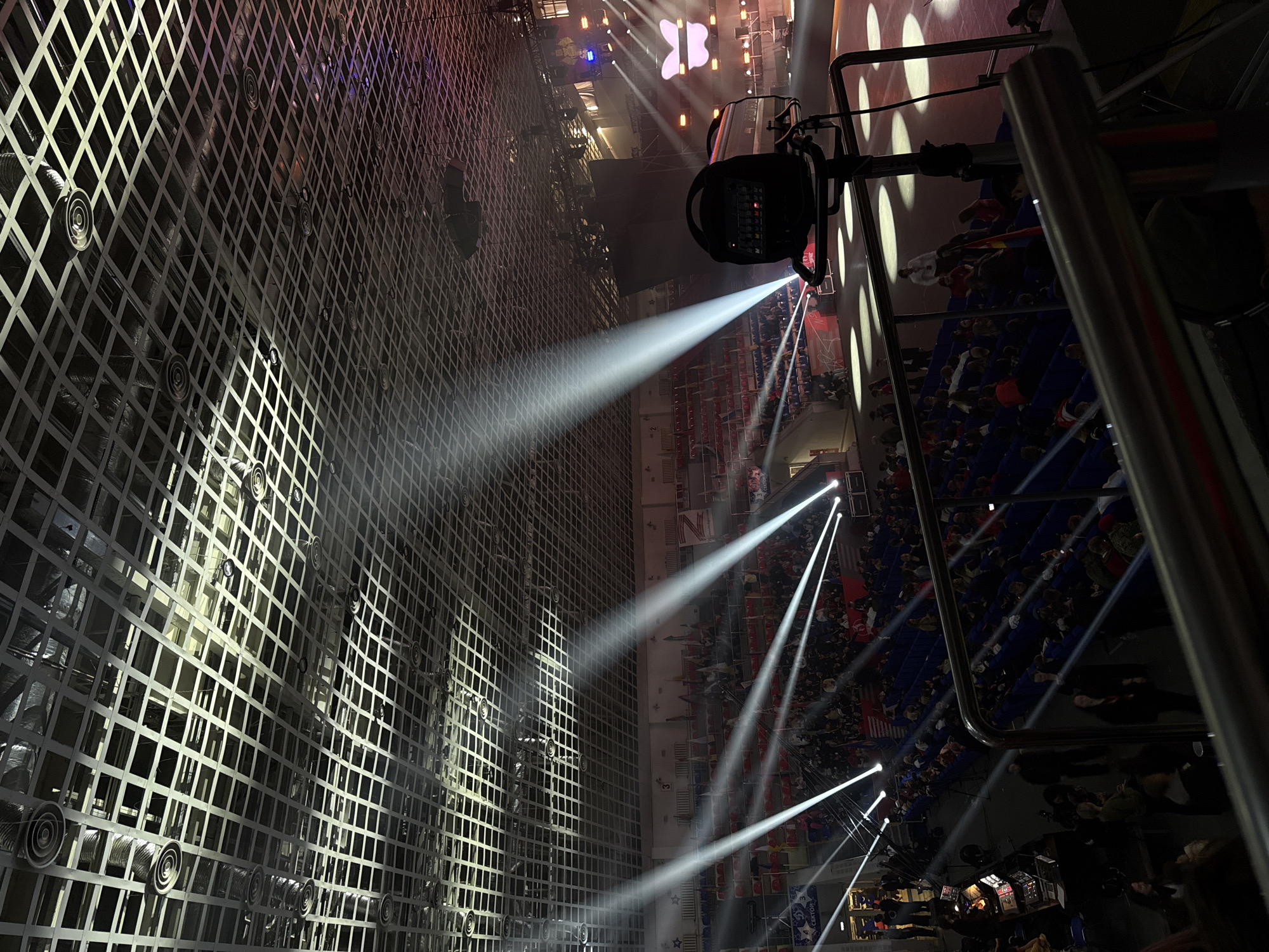 The image depicts an indoor venue with a large crowd seated in tiered rows. The venue appears to be a stadium or arena, possibly for a sporting event or concert. Stage lighting is evident, with spotlights shining beams across the room. A grid-like structure occupies a significant portion of the left side of the image.