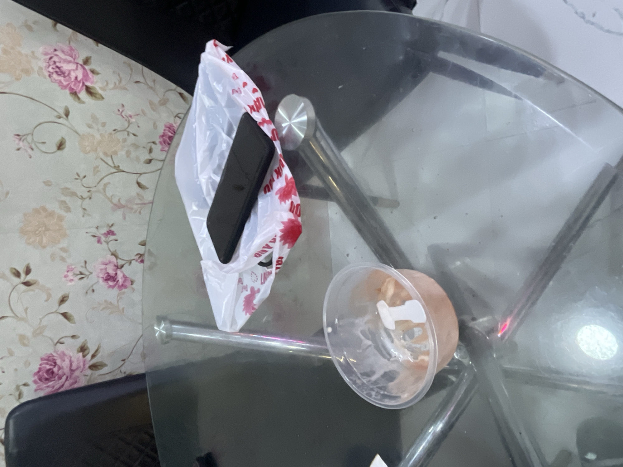 An overhead shot shows a glass-top table with a phone, a plastic takeout container, and a plastic bag on it. The table is in a room with floral wallpaper and two dark chairs. The scene is illuminated by overhead lighting, creating reflections on the glass surface. The takeout container appears to have some residue inside, and the plastic bag, which is partially open, contains the phone and has red and white floral designs printed on it. The phone is black and rectangular, placed on top of the bag. The environment suggests a casual indoor setting, possibly a dining area. No specific location in Jalingo, Nigeria can be determined from the image.
