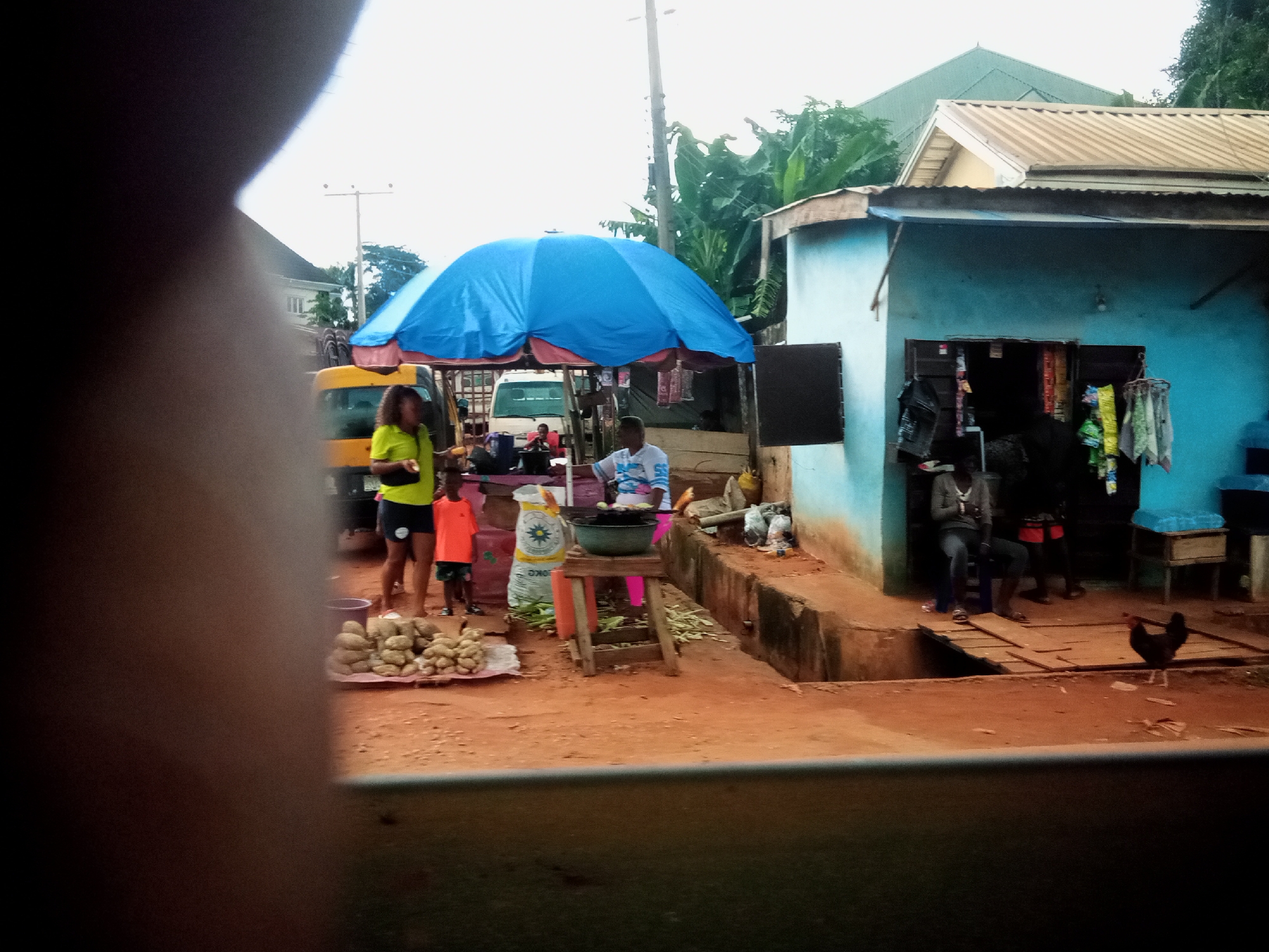 The image depicts a bustling outdoor market scene in Nibo, Nigeria, on an overcast day. In the foreground, a partially visible person's arm obscures part of the view. The central area features a bright blue and red umbrella, under which a woman in a green shirt and dark shorts is standing near a yellow vehicle. Beside her, a young boy in an orange shirt and shorts stands next to a pile of produce, likely yams, arranged on a mat. A large white bag with a green logo is also present. Further to the right, a blue building serves as a storefront with open doorways displaying various goods. A person in a grey top and dark pants sits on a stool outside the building, and another person is visible inside. A wooden structure with a shallow trench in front of it connects the market stalls. A dark-colored chicken is walking on the reddish-brown dirt ground in the lower right corner. The background shows a residential structure with a corrugated metal roof and trees. The overall impression is one of daily life and commerce in a Nigerian town.