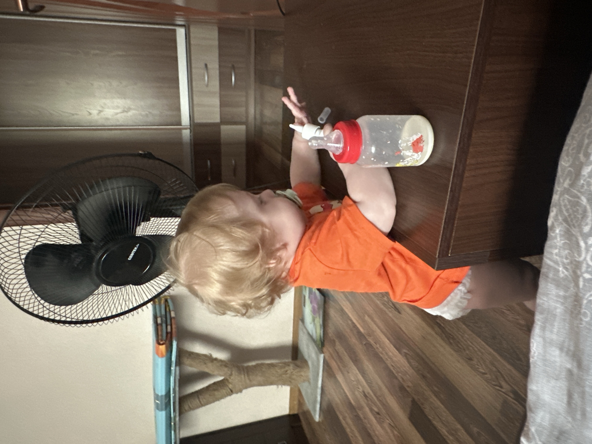 A toddler with blond hair is standing next to a dark wooden table, reaching for a baby bottle with a red cap. The toddler is wearing an orange t-shirt and a diaper with white lace trim. A pacifier is in the toddler's mouth. In the background, a black fan is visible, along with a cat tree and wooden cabinets. The scene appears to be indoors, in a residential setting in Sayanogorsk, Russia.