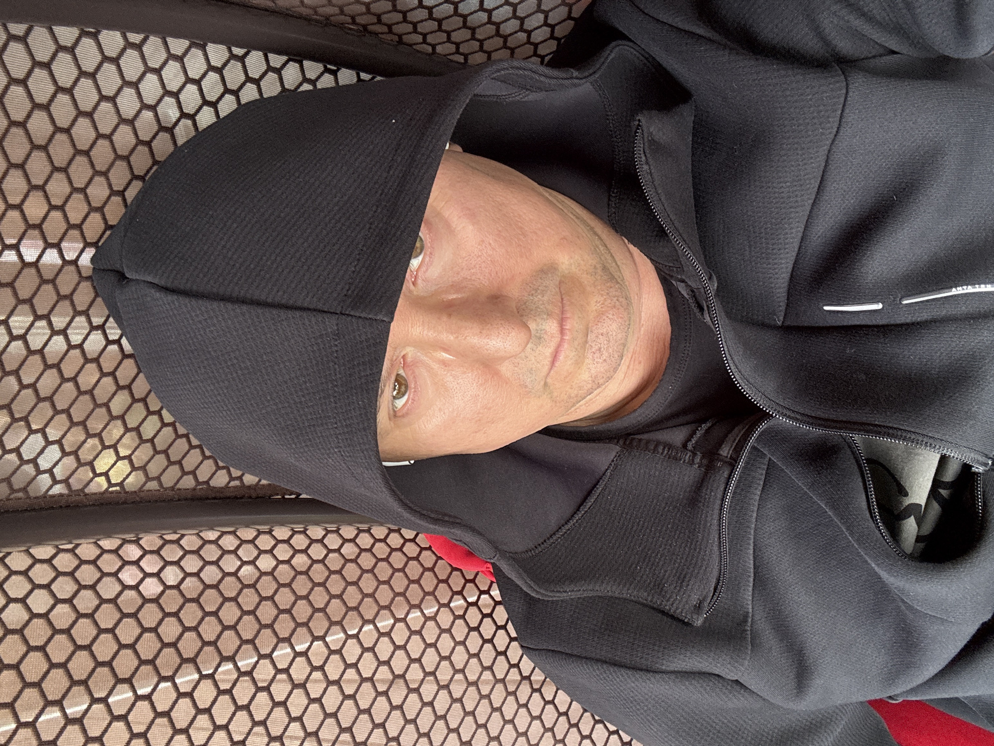 A man wearing a black hooded jacket is shown in a close-up, selfie-style photograph. The background consists of a textured, honeycomb-patterned mesh in a reddish-brown color. The man's face is partially obscured by the hood of his jacket. He has light skin, brown eyes, and is looking directly at the camera with a neutral expression. The image does not provide any information about a location in Gavshino, Russia.