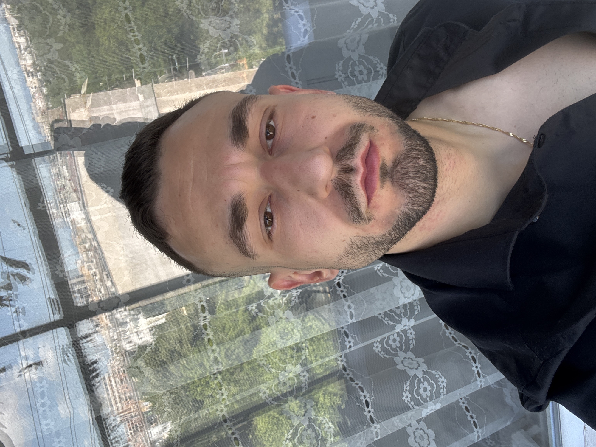 A selfie-style photograph shows a man with short, dark hair, light brown eyes, and a trimmed beard. He is wearing a black shirt and a thin gold chain around his neck. The background reveals a window with a sheer, patterned curtain, partially obscuring a view of what appears to be a city.
Based on visual clues such as architectural styles, and the overall urban landscape visible through the window, and in conjunction with the user question, it can be inferred that this photo could have been taken in Plovdiv, Bulgaria. The photograph shows buildings and greenery indicating the location is an urban area. However, precise identification of a specific location within Plovdiv is not possible with the provided image.